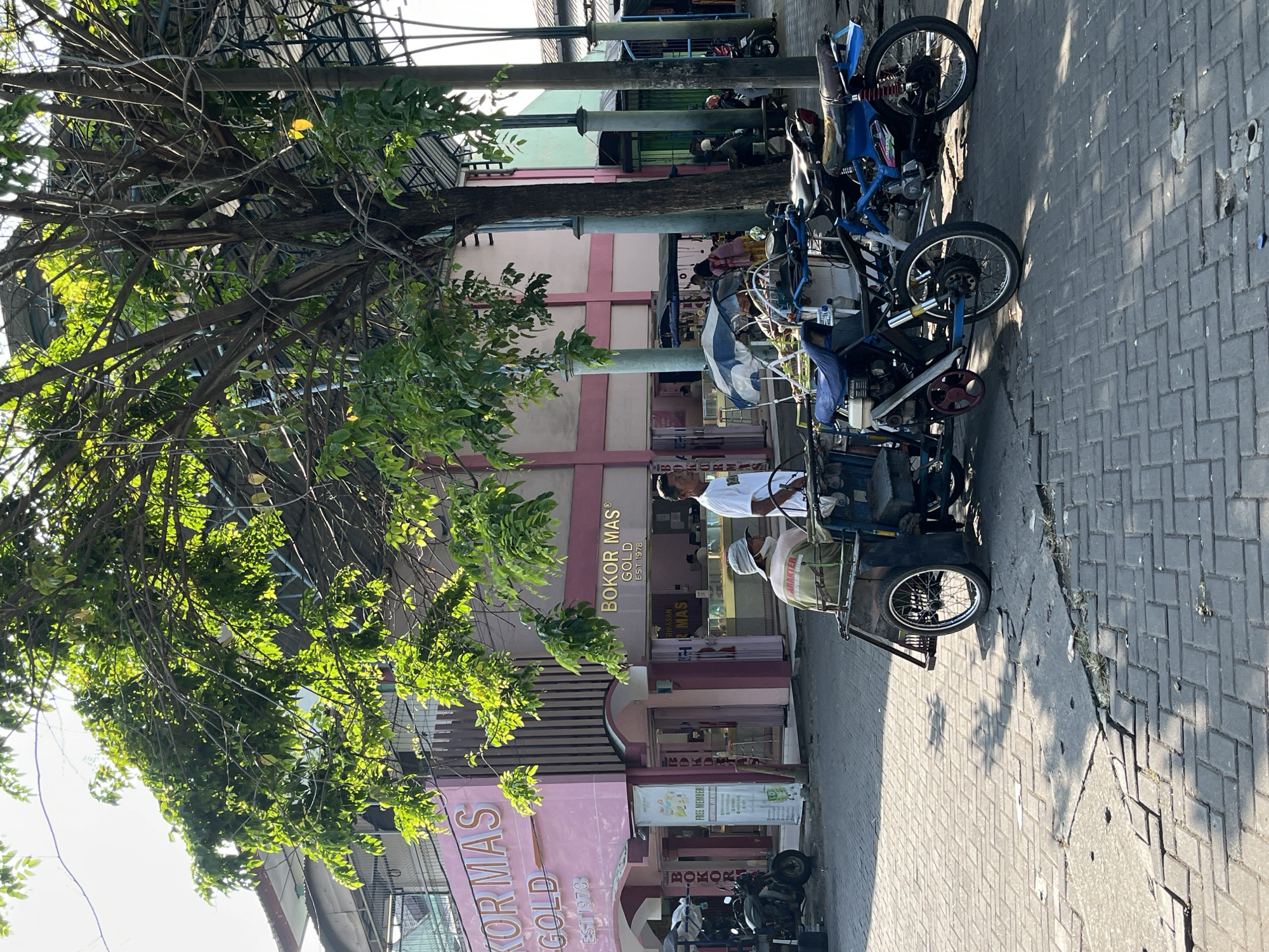 This image depicts a street scene in Juwet Kenongo, Indonesia. The foreground is dominated by a paved street with visible cracks and uneven sections. A three-wheeled vehicle, possibly a modified motorcycle or a small cart, is positioned in the lower center, featuring a blue fabric cover and a person wearing a light-colored hat and a green garment seated on it. Next to it, a blue motorcycle is parked.
In the middle ground, two men are standing near the three-wheeled vehicle. One man, wearing a white shirt, appears to be interacting with the other man who is seated. Behind them, a pink building with "BOKOR MAS GOLD EST 1978" signage is visible. The building has a striped facade and several entrances. A vertical sign with text and graphics is also displayed on the building's exterior. Several motorcycles are parked in front of the building.
The background reveals more of the street and buildings, with some trees providing shade. The overall atmosphere suggests a commercial area, likely a market or a street with shops. The lighting indicates it is daytime, with sunlight casting shadows on the pavement.