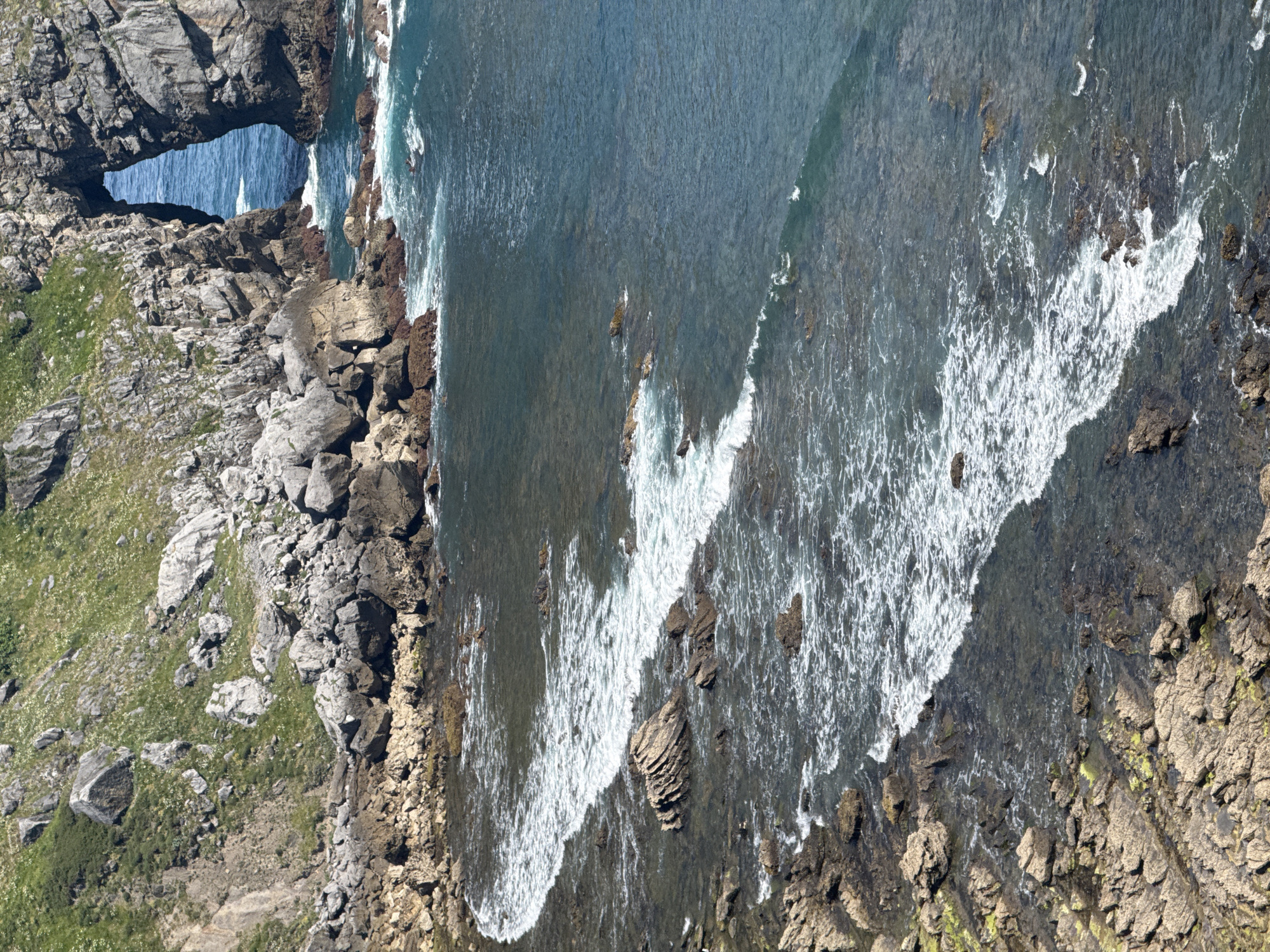 This is a high-angle, long shot of a rocky coastline in Bermeo, Spain. The image captures the dynamic interplay between land and sea, with waves crashing against the shore. The foreground is dominated by rough, textured rocks, some covered in a light green moss or algae. Interspersed among these rocks are patches of shallow, dark water. Moving further back, a white-capped wave is seen rolling towards the coast, its foamy crest indicating its momentum. Beyond this, the water becomes deeper and a richer blue. In the background, a prominent natural archway carved into a cliff face is visible. This archway frames a view of the bright blue sea. The cliff face itself is steep and rocky, with patches of green vegetation growing on its upper slopes. The overall impression is one of a rugged and untamed natural landscape.