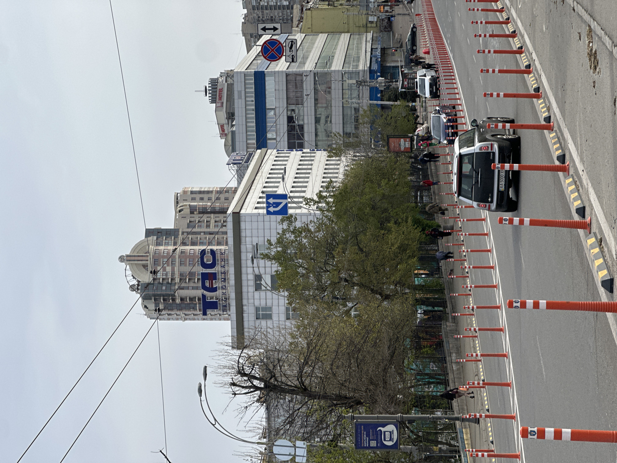 An urban street scene in Kyiv, Ukraine, is depicted on what appears to be a bright, overcast day. The view looks down a street with buildings lining the left side and a multi-lane road on the right, separated by a continuous line of orange and white traffic bollards.
On the left, several multi-story buildings are visible. A prominent white building in the mid-ground features a large blue vertical logo spelling "ТЭС" (TES) with "ТАСКОМБАНК" (TASKOMBANK) written horizontally beneath it, indicating a bank. Further down, another building has "HOTEL" signs on its upper facade. A distinct building with a dome-like roof stands in the background. Various trees with mixed foliage, some bare and some with green leaves, line the street. Overhead power lines crisscross the sky.
The road on the right has several vehicles, including a black mini-van or small SUV with the license plate "AA 5000 XX" in the lane closest to the bollards. Other cars are visible further up the street. Several pedestrians are scattered along the sidewalk and near the road.
Notable street furniture and signs include a blue and white "bus stop" sign with a QR code, a red circular "no stopping" or "no parking" sign, a blue directional sign with white arrows, and another sign indicating "no entry" or "no vehicles." Streetlights are also present. The sky is bright and uniformly overcast.