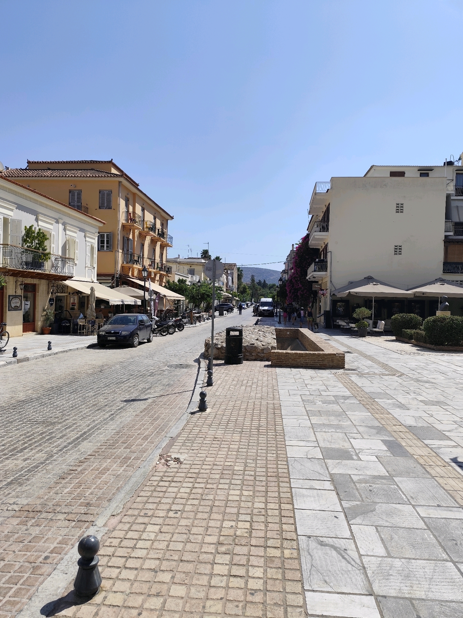 The image depicts a street scene in the Municipal Unit of Nafplio, Greece, on a clear, sunny day. The street is paved with cobblestones, and there is a sidewalk made of large, light-colored tiles on the right. Buildings line both sides of the street. On the left, a multi-story building with a yellow facade features balconies with ornate railings. Below, awnings extend over storefronts, suggesting commercial establishments such as cafes or shops. A dark-colored car is parked on the street. On the right, a white multi-story building has a balcony and flowering vines. Further down the street on the right, there are outdoor seating areas under umbrellas, indicating a cafe or restaurant. People are visible walking on the sidewalk on the right. In the distance, hills or mountains are faintly visible. The sky is clear blue. Several black bollards line the edge of the cobblestone street and the sidewalk. A trash can is situated near a stone and brick structure on the right sidewalk. The overall impression is of a well-maintained urban environment in a Mediterranean setting.