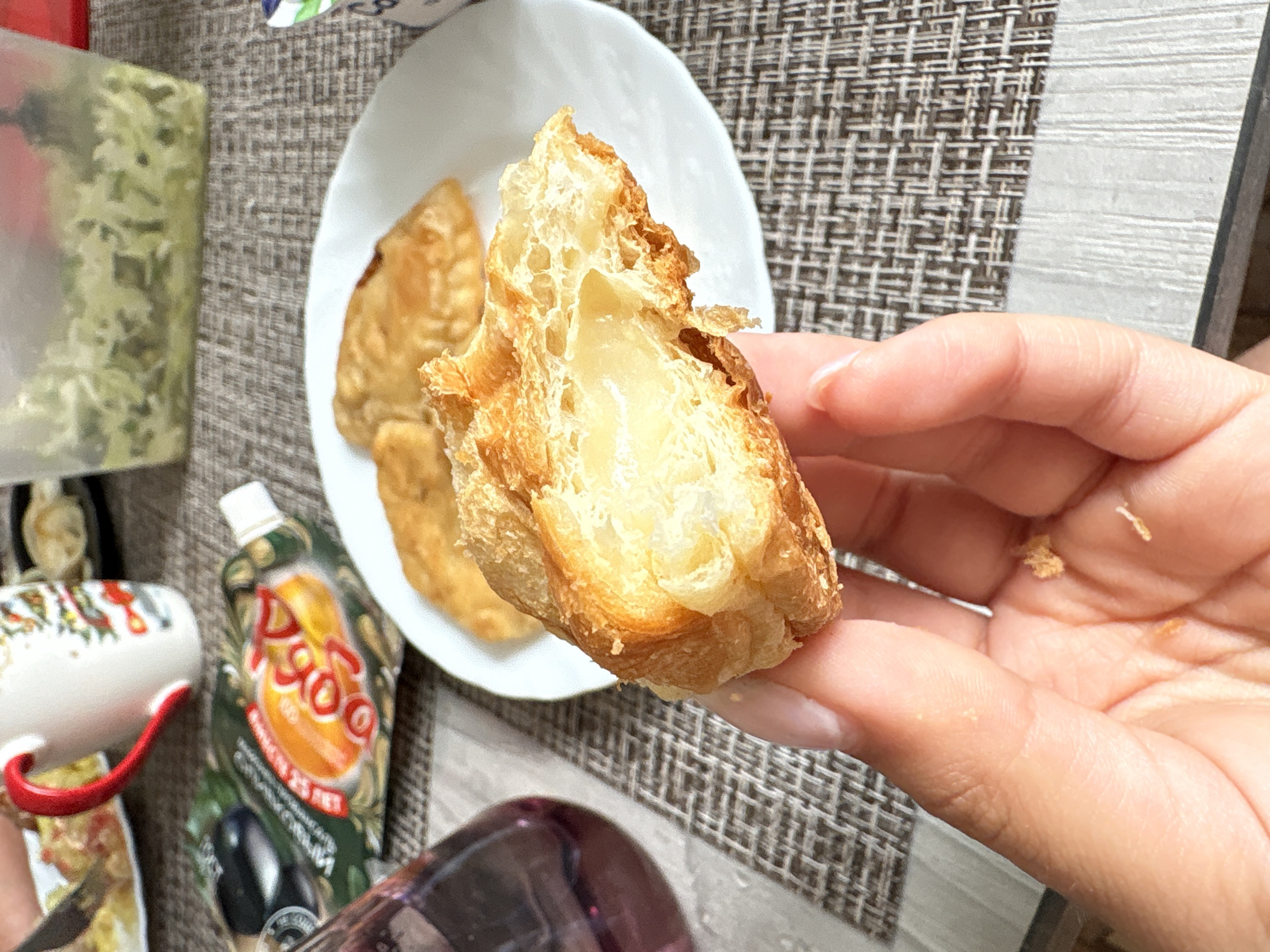 A close-up indoor shot captures a person's hand holding a broken piece of golden-brown, flaky pastry, revealing a creamy, light-colored filling inside. The pastry appears to be a type of puff pastry or croissant. In the background, on a white plate, are at least two other similar pastries. The scene is set on a patterned placemat, featuring a grid-like design in grey and white tones.
Various objects are partially visible around the main subject. To the left, a transparent container holds light green, shredded contents, possibly herbs or a finely chopped salad. Below this, a white mug with a red handle and festive red and green patterns can be seen. A squeeze pouch of a food product, possibly a sauce or mayonnaise, with a prominent "РОДО" or "ДОДО" brand logo in orange Cyrillic text, is visible. The pouch is green and depicts an olive-like fruit. Visible Cyrillic text on the pouch includes "Оливки 25 лет" (Olives 25 years) and "Оливковый соус" (Olive sauce). A dark-colored glass or bottle is also partly seen in the lower right corner. The lighting suggests daytime. The visible Cyrillic text on the food packaging is a strong indicator of the location being a Russian-speaking region, consistent with the Прикамский, Russia context.