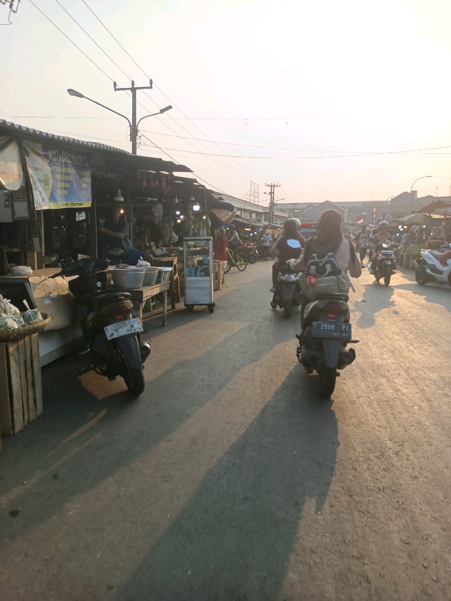 The image depicts a busy street market in Karawang, Indonesia, during what appears to be late afternoon, with the sun casting long shadows. On the left side of the road are market stalls with awnings. One stall displays a banner with Indonesian text and a logo. Various goods are visible on display, including what looks like produce and household items. Several motorcycles are parked in front of or near the stalls. A man is seated at one of the stalls, seemingly attending to business.
The road itself is paved and shows signs of wear. Traffic is present, with several people riding motorcycles. In the foreground and mid-ground, motorcycles are the dominant moving subjects. One motorcycle in the mid-ground has a rider wearing a hijab and a backpack, and another motorcycle behind it also has a rider. Further down the road, more motorcycles and people are visible, suggesting a lively commercial environment. The sky is bright, indicating daylight, and there are power lines and streetlights visible above the market stalls. The overall scene conveys a sense of everyday activity and commerce in a semi-urban Indonesian setting.