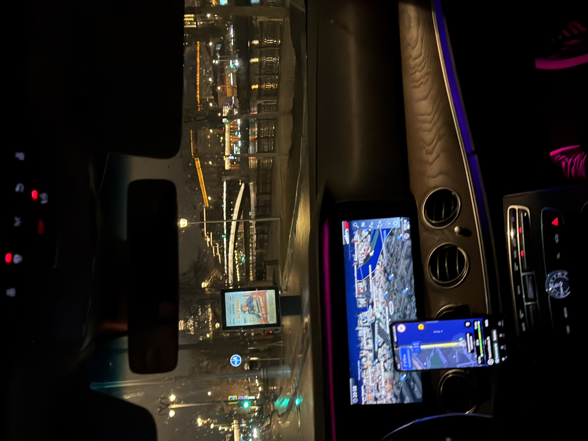 The image is taken from inside a car at night, likely in Moscow, Russia. Outside the car, a city street is visible with lights, buildings, and wet road reflecting the light. An advertising billboard is on the street, featuring what appears to be a movie poster titled "Al RAMO". Inside the car, the dashboard is visible. A large infotainment screen shows a map, presumably for navigation, and a smartphone is mounted near it, also displaying a navigation app. Part of the steering wheel and the center console are visible, with controls and air vents. The interior is dark, but the ambient light illuminates some of the car's features. The overall impression is of a car traveling through a city at night, guided by navigation systems.