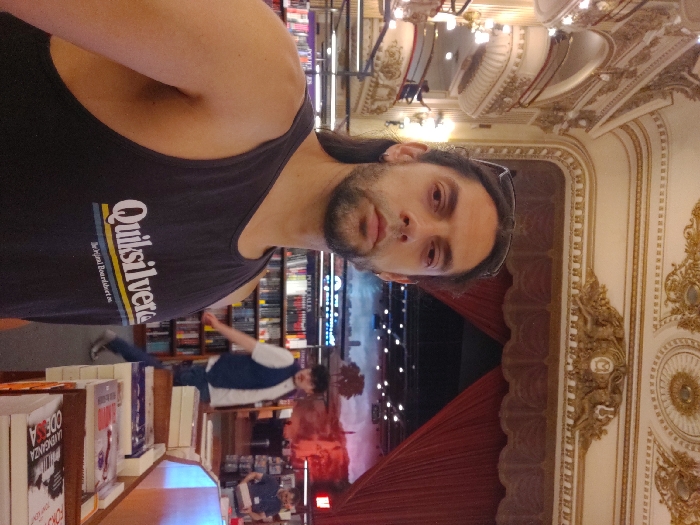 A man wearing a black tank top with "Quiksilver" printed on it poses in what appears to be a bookstore or theater. Books are visible on shelves in the background. Further back, there is a person in a vest and a performance on a stage. The ornate architecture and design of the building are also apparent. The building's style suggests it may be a historical theater.