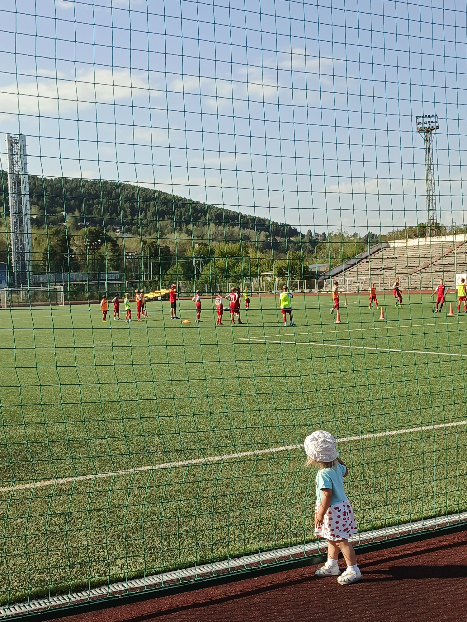 The image depicts a children's soccer practice at a stadium in Zlatoust, Russia. In the foreground, a young girl in a blue shirt and a white hat with a cherry-patterned skirt, stands facing away from the camera, looking towards the soccer field. She is wearing white sandals and white socks. A green net separates her from the field. On the field, several children in red and white soccer uniforms are engaged in drills, with orange cones visible. In the background, stadium seating, trees, hills, and a transmission tower are visible under a bright sky with scattered clouds. The scene suggests a sunny day at an outdoor sports facility.