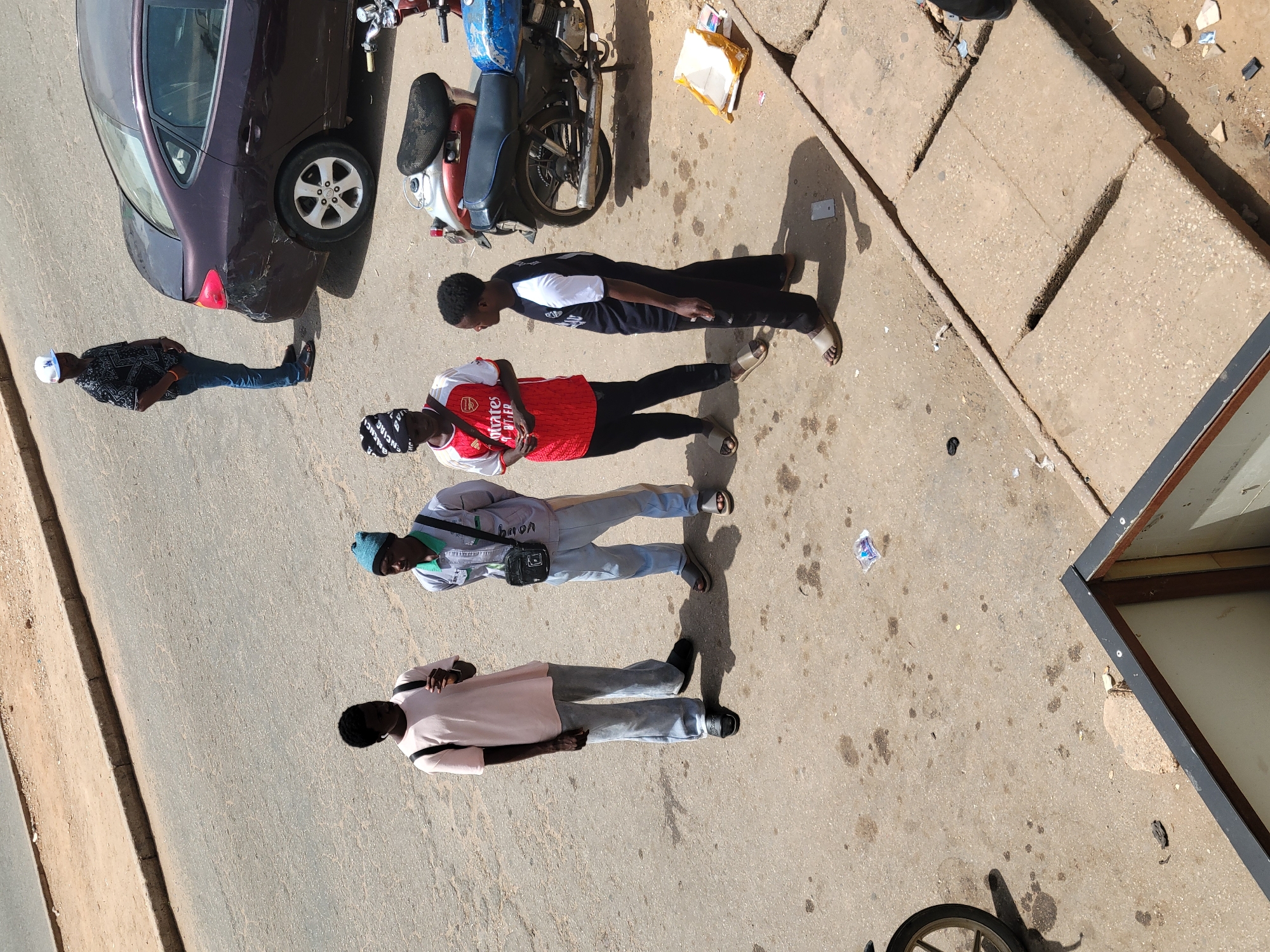 The image, taken from an elevated perspective, shows an outdoor scene in the city of Jos, Nigeria. Four individuals are standing on a paved surface, likely a street or sidewalk. One person is wearing a patterned shirt and jeans with a white cap, while another is in a red Arsenal jersey, black pants, and a black patterned head covering. A third individual is wearing a light blue jacket, light grey pants, and a blue beanie. The fourth person is dressed in a light pink shirt and grey pants.
A dark purple car is parked on the left side of the frame. A motorcycle is parked near the car. Debris and litter are scattered on the ground. The right side of the image shows a section of a building with a metal frame.