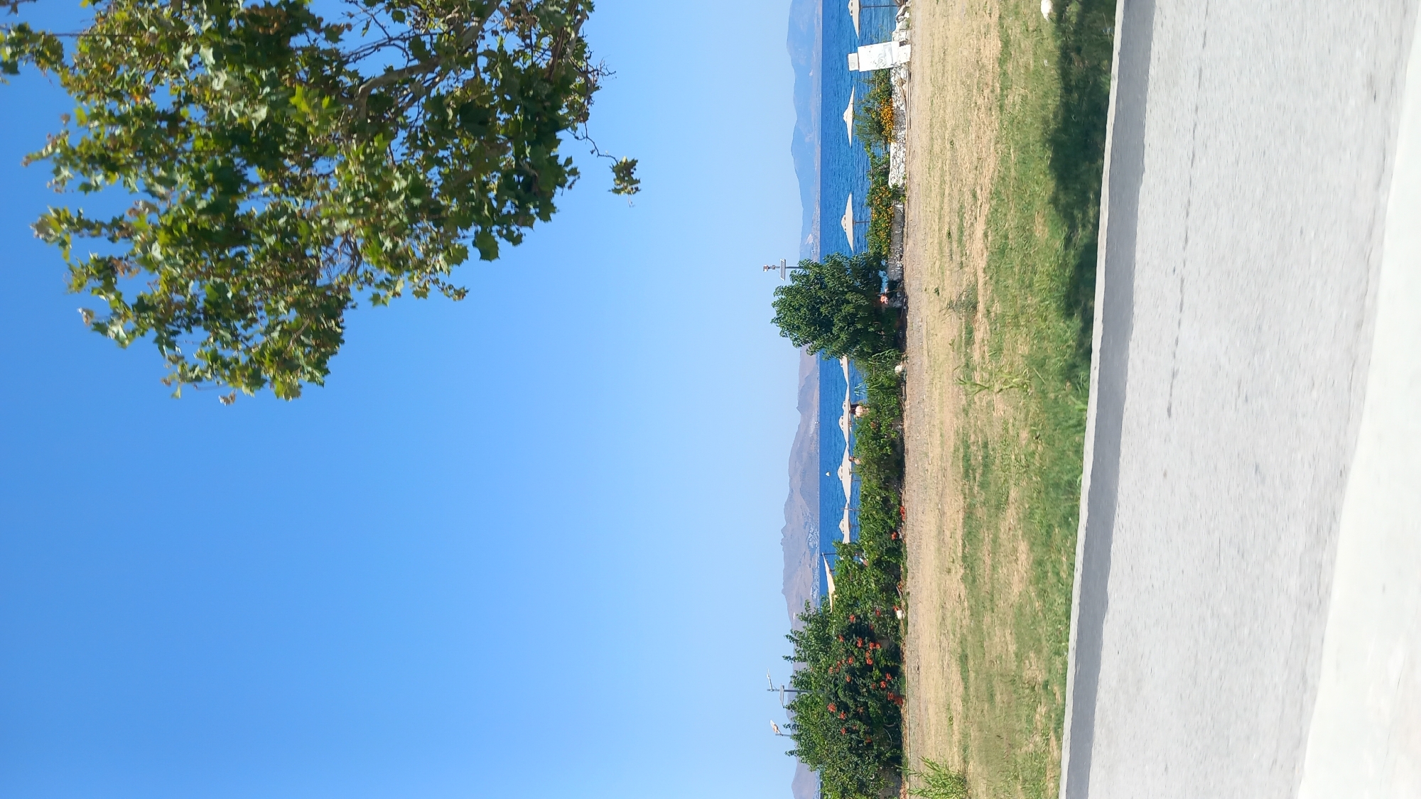 The image depicts a scenic view of a beach and the sea. In the foreground, there is a paved road and a grassy area. Beyond the grass, there is a line of beach umbrellas and some vegetation, including bushes and trees. The sea is visible in the background, with mountains on the horizon. The sky is clear and blue. The scene appears to be a sunny day at a coastal location.