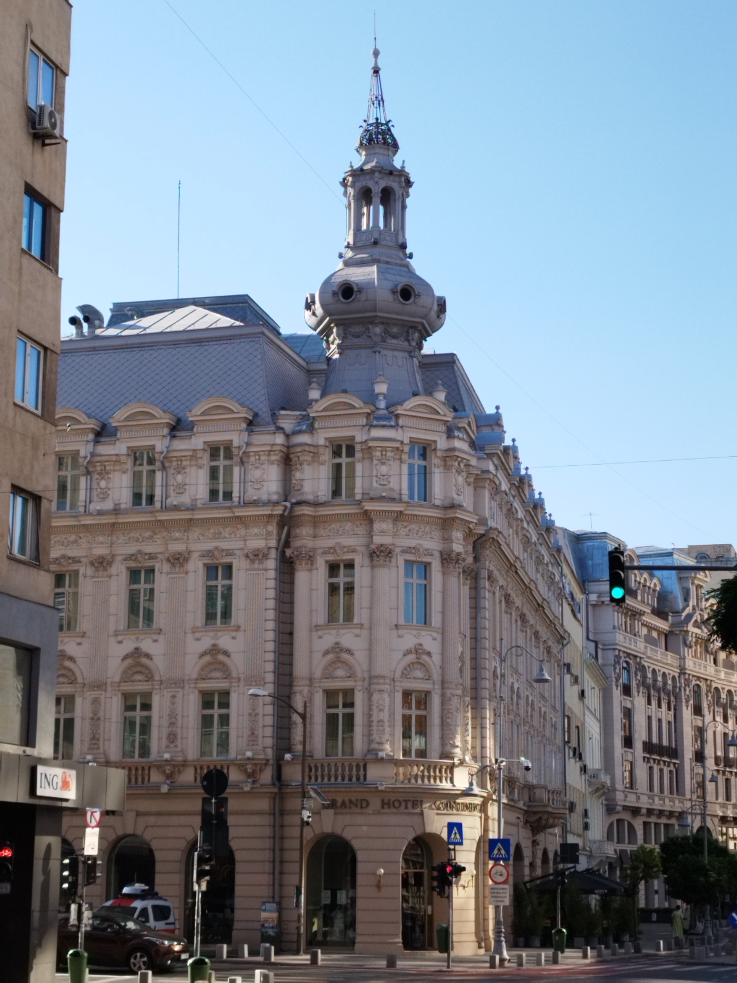 This is a daytime, eye-level, medium-shot photograph of a street scene in Bucharest, Romania, featuring ornate architecture. On the left is a lighter-colored, older building with multiple windows and an air conditioning unit. The central and right portions of the image are dominated by a grand, multi-story building with intricate detailing, ornate window frames, and a distinctive domed tower topped with a spire. This building appears to be the Grand Hotel Continental, as suggested by visible signage. The facade is light-colored, possibly stone or stucco, with numerous windows across its levels. The building has a mansard roof with dormer windows. A green traffic light is visible on the right side of the frame, indicating an intersection. The street is paved, and there are traffic signals, streetlights, and bollards. A dark-colored car is parked on the right side of the street. The sky is clear and blue.
