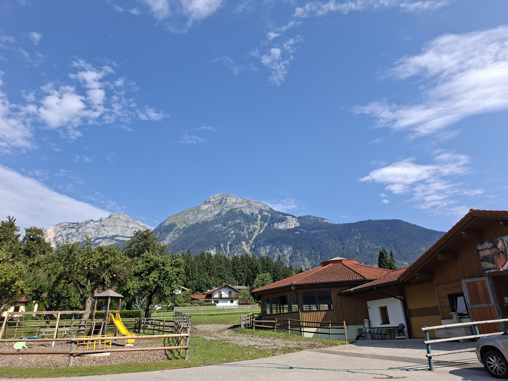 This is a horizontal, outdoor, medium-shot photograph taken on a sunny day in Münster, Austria. The scene depicts a rural setting with a playground and buildings against a backdrop of majestic mountains.
In the foreground, a wooden fence encloses a playground with a yellow slide, a wooden climbing structure, and a swing. To the right of the playground, a wooden building with a red-tiled roof and large windows is visible. A car is parked to the far right of the frame. Further back, behind the playground and the first building, are more wooden fences and green fields. Several trees, some laden with fruit, are scattered throughout the scene.
In the mid-ground, a cluster of houses with white and brown exteriors nestle amongst trees. Dominating the background is a vast, rugged mountain range. The lower slopes of the mountains are covered in dense, dark green conifer forests, while the upper reaches are characterized by rocky, grey cliffs and peaks. The sky above is a brilliant blue, sparsely dotted with white, fluffy clouds.
The overall impression is one of a peaceful, natural landscape complemented by recreational facilities and rustic architecture, characteristic of an Austrian rural resort or farm. The image captures a tranquil, idyllic moment in the Austrian countryside.