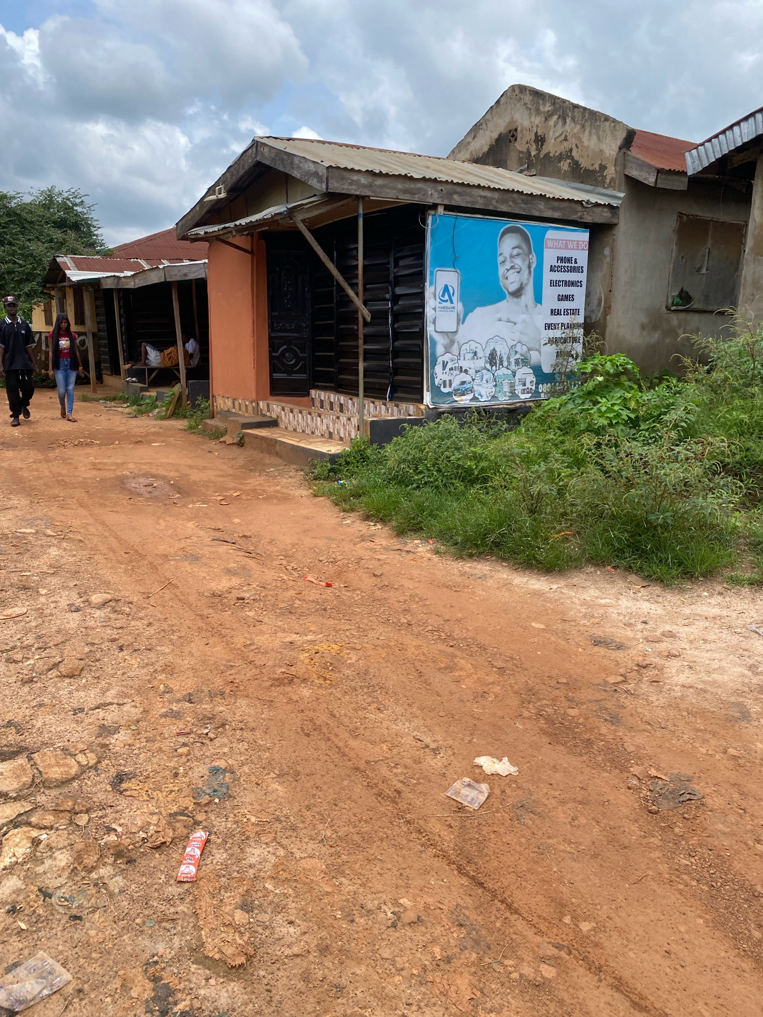In Iworoko, Nigeria, a dusty, red-dirt road leads past a row of small shops with corrugated metal roofs. Two individuals, a man in a black shirt and cap and a woman in a red shirt and blue jeans, walk along the road towards the left of the frame. A person is seen resting on a bench outside one of the shops. A large blue and white advertisement poster is attached to the front of one shop, listing services like "Phone & Accessories," "Electronics," "Games," "Real Estate," "Event Planning," and "Agriculture." The environment is characterized by overgrown green vegetation encroaching on the road and buildings. The ground is littered with debris, including plastic wrappers and other refuse, suggesting a lived-in and perhaps less-than-pristine urban setting. The sky is overcast with clouds.