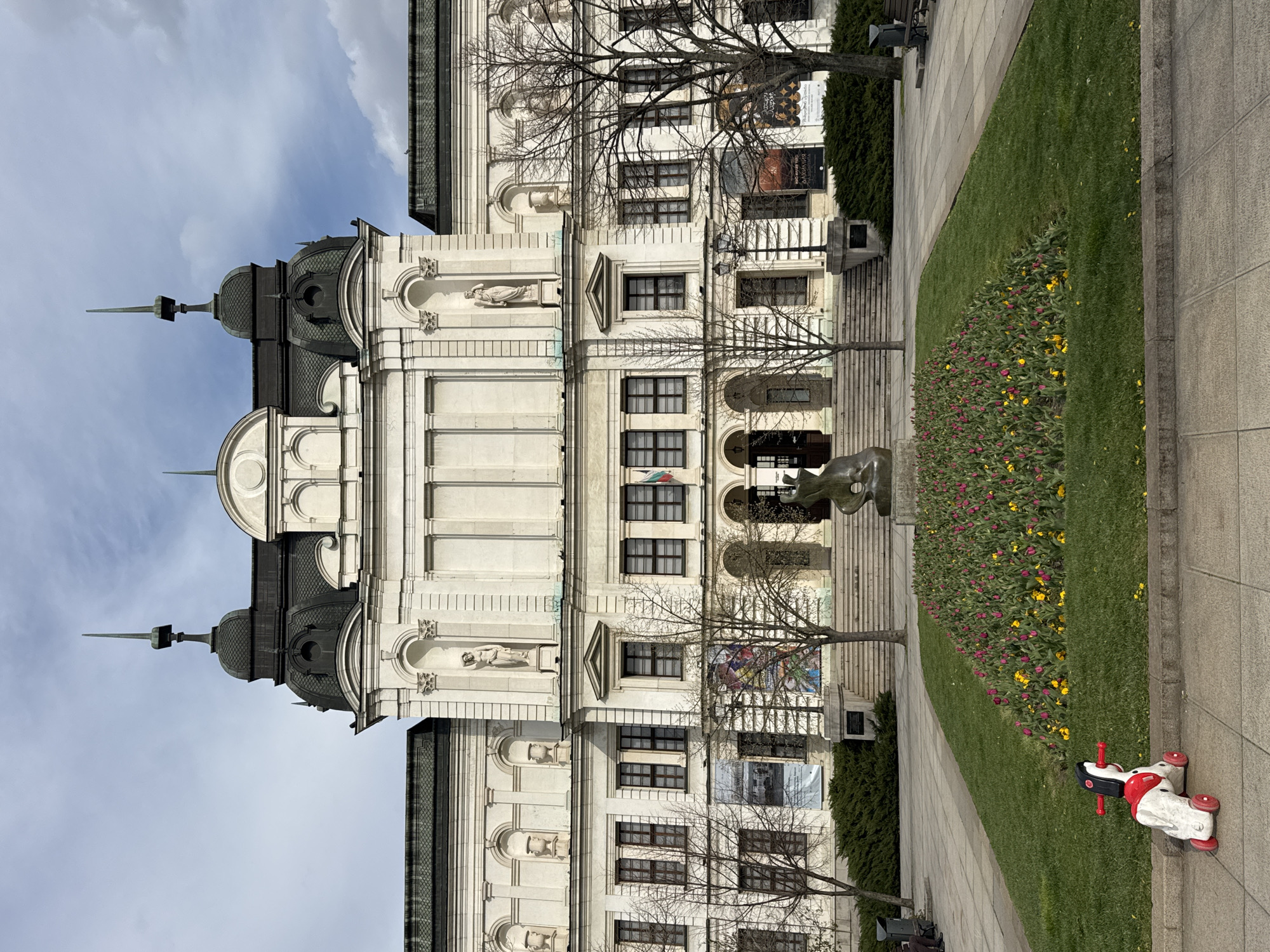 The image shows a building in Sofia, Bulgaria, likely the National Art Gallery. It's a multi-story, white building with decorative elements like statues and archways. There are trees with bare branches in front of the building. A green lawn with a flowerbed containing tulips stretches along the right side of the frame. A sculpture is also visible. On the sidewalk, a child's ride-on toy is present. The sky is partially cloudy.