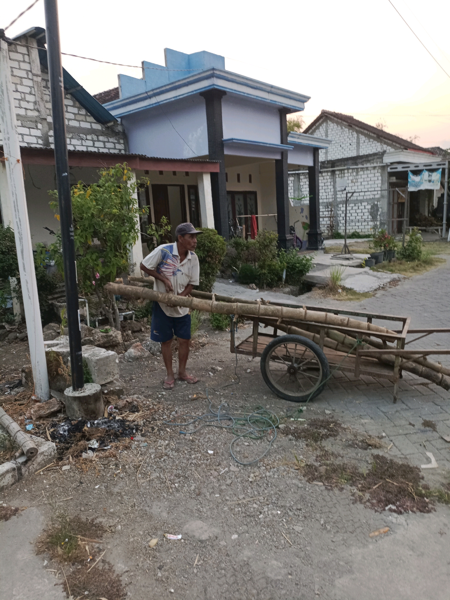 In Rancang Kencono, Indonesia, a man in a light-colored shirt and dark shorts stands outdoors next to a two-wheeled cart. He is holding a long bamboo pole horizontally across his body, with one end resting on the ground and the other supported by his left arm. The cart, made of metal and wood, contains several more bamboo poles. The ground is a mix of gravel, dirt, and some concrete. Several houses are visible in the background, featuring white brick walls and pitched roofs. Vegetation, including bushes and trees, is scattered around the scene. A utility pole stands to the left of the man. The sky suggests it is either early morning or late afternoon.