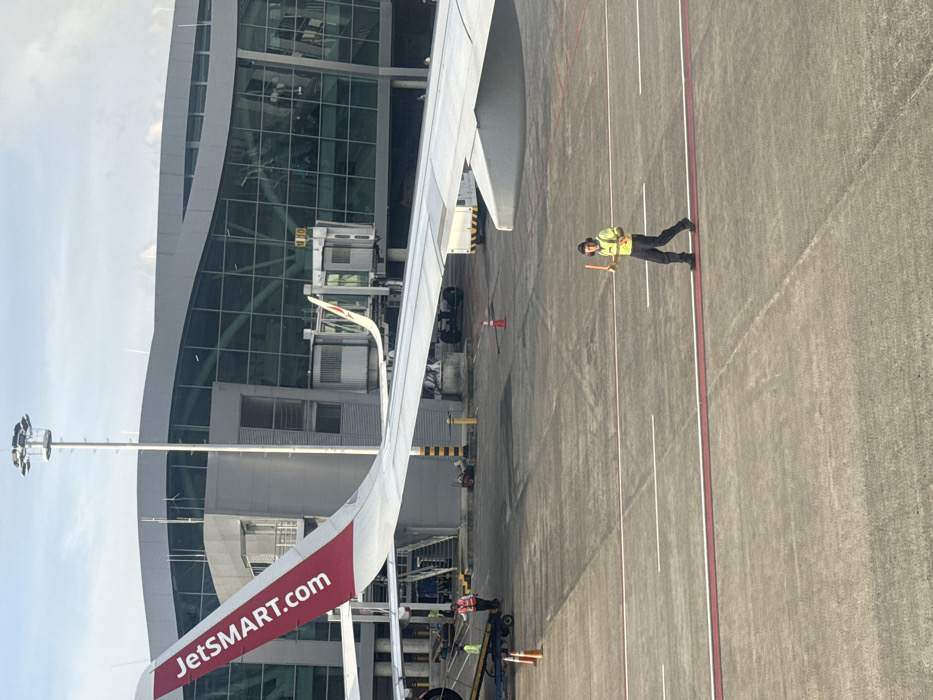 An airport apron view shows a JetSMART plane wing with the company's website prominently displayed. A ground crew member, wearing a safety vest and holding a signal wand, stands beside the wing. The modern airport terminal, featuring a curved glass facade, is visible. The ground is marked with painted lines and tire tracks, illustrating an active airfield environment.