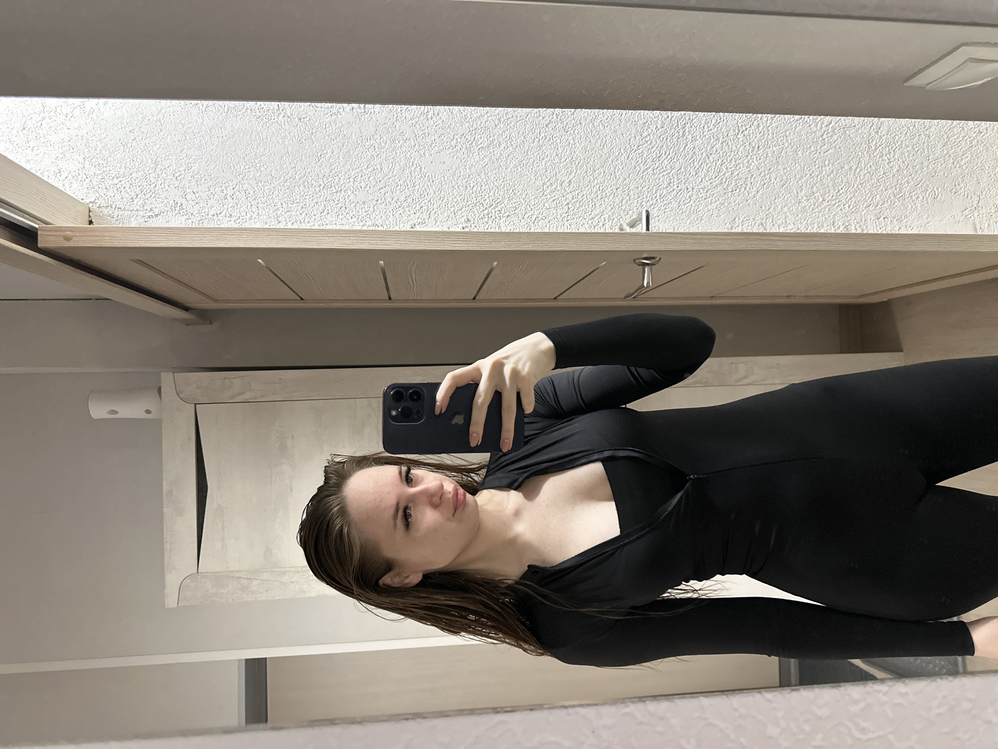 A full-length shot features a woman taking a selfie in what appears to be a bathroom. She is wearing a black, fitted bodysuit and is holding a black smartphone. Her wet hair is combed back, and she looks directly at the camera. The background includes a light-colored cabinet with a textured white wall above it. A small metal hook is visible on the cabinet. The location is in the city of Unknown, Unknown.