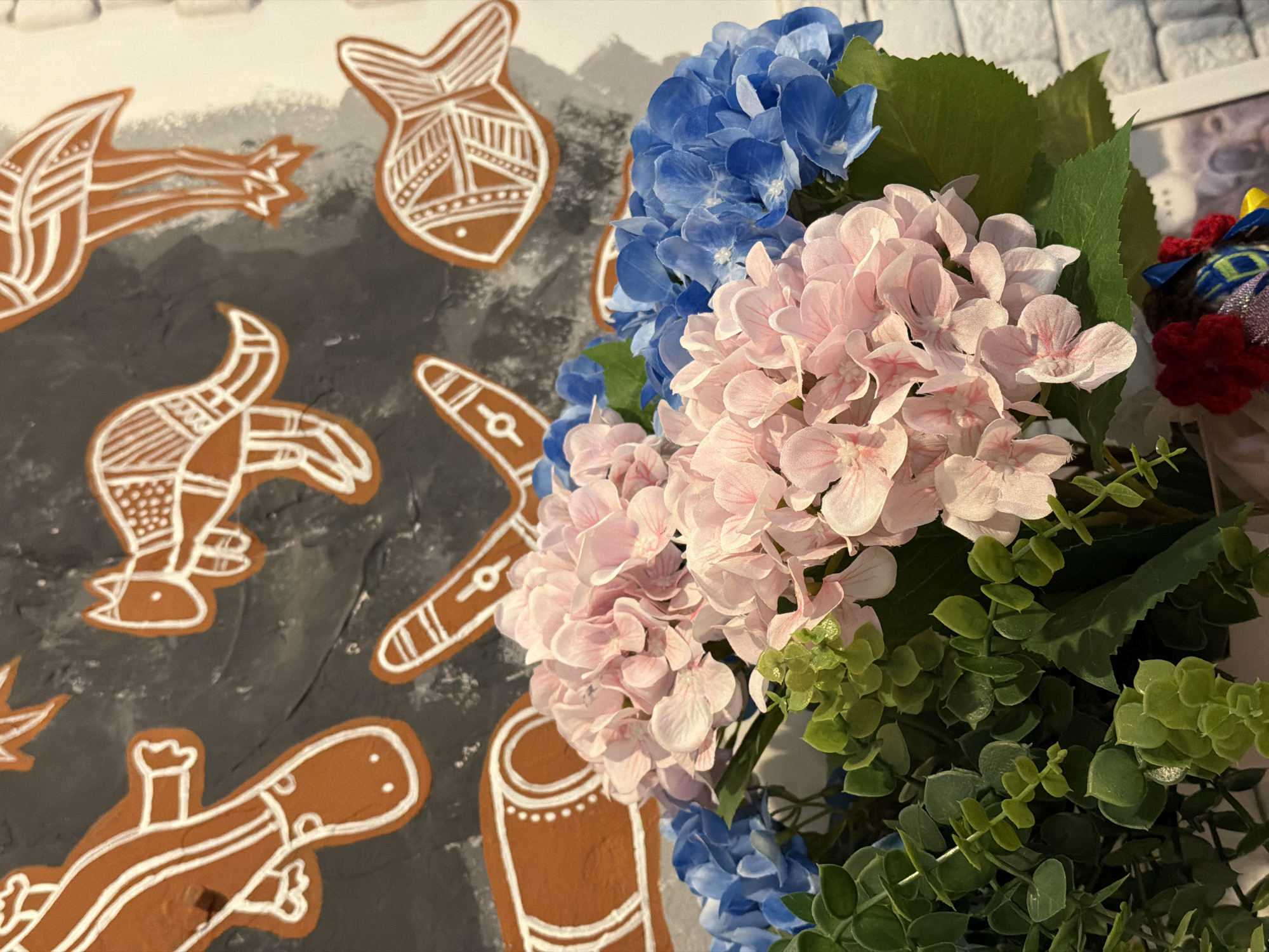 The image shows a close-up of an arrangement of blue and pink hydrangeas with green foliage, positioned in front of a painted wall. The wall features brown and white depictions of stylized animals and objects on a grey and white background. The animal/object depictions include a lizard, fish, boomerang, and other abstract forms. The location is not clearly visible in the provided image to confirm it is in Dnipro, Ukraine.