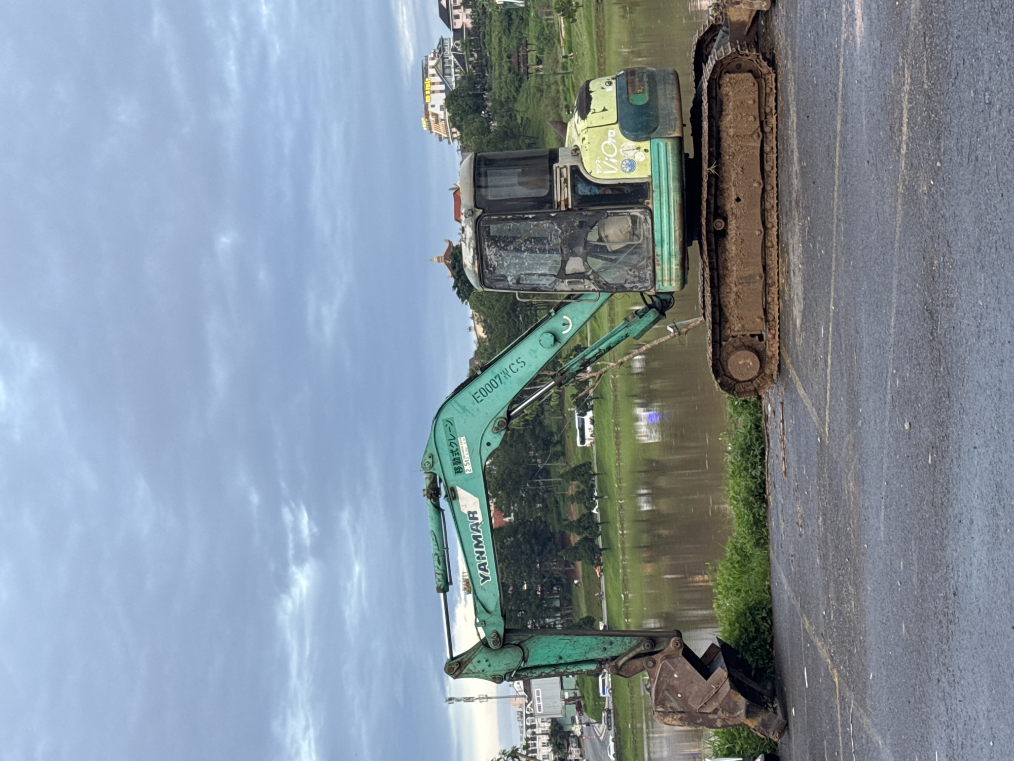 The image shows a green and beige Yanmar excavator parked on a grey asphalt surface, possibly a parking lot, with its tracks caked in mud. The excavator's arm extends towards a body of water, reflecting the sky and surrounding trees. In the background, there is a view of buildings nestled among the trees, typical of the architecture of Da Lat, Vietnam. The sky is overcast with grey clouds. The excavator is located next to a lake.
