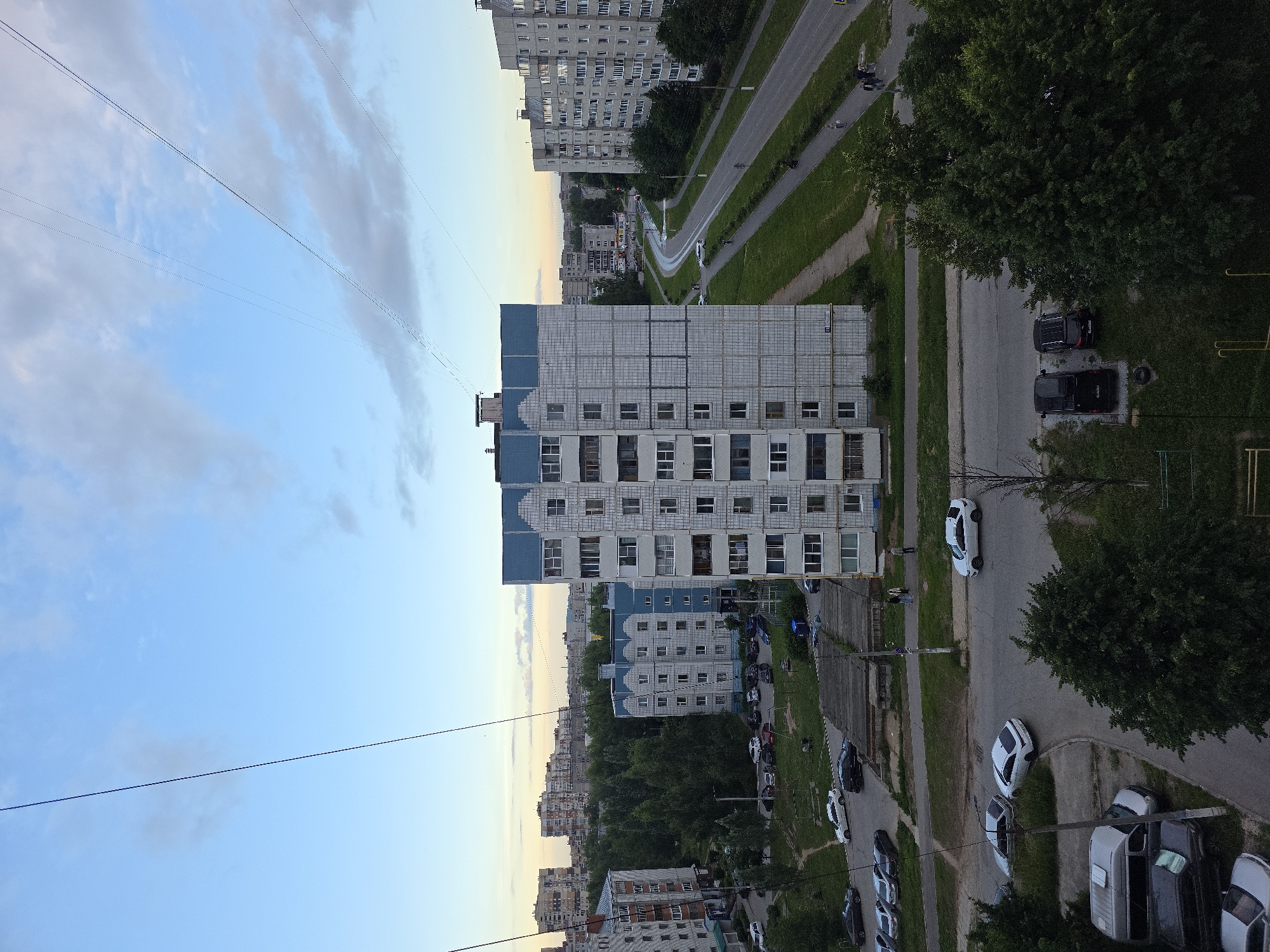 The image shows an aerial view of a residential area in Novocheboksarsk, Russia. The scene is dominated by multi-story apartment buildings, some with blue accents on their upper floors and rooftops. Utility wires crisscross the sky above the buildings. A paved road runs through the scene, with cars parked along its sides and some driving on it. Green spaces, including trees and grassy areas, are interspersed between the buildings and along the road. A few people are visible walking on sidewalks and in grassy areas. The sky is partly cloudy with a soft, diffused light, suggesting it may be evening or early morning. The architecture appears to be typical of Soviet-era residential developments.