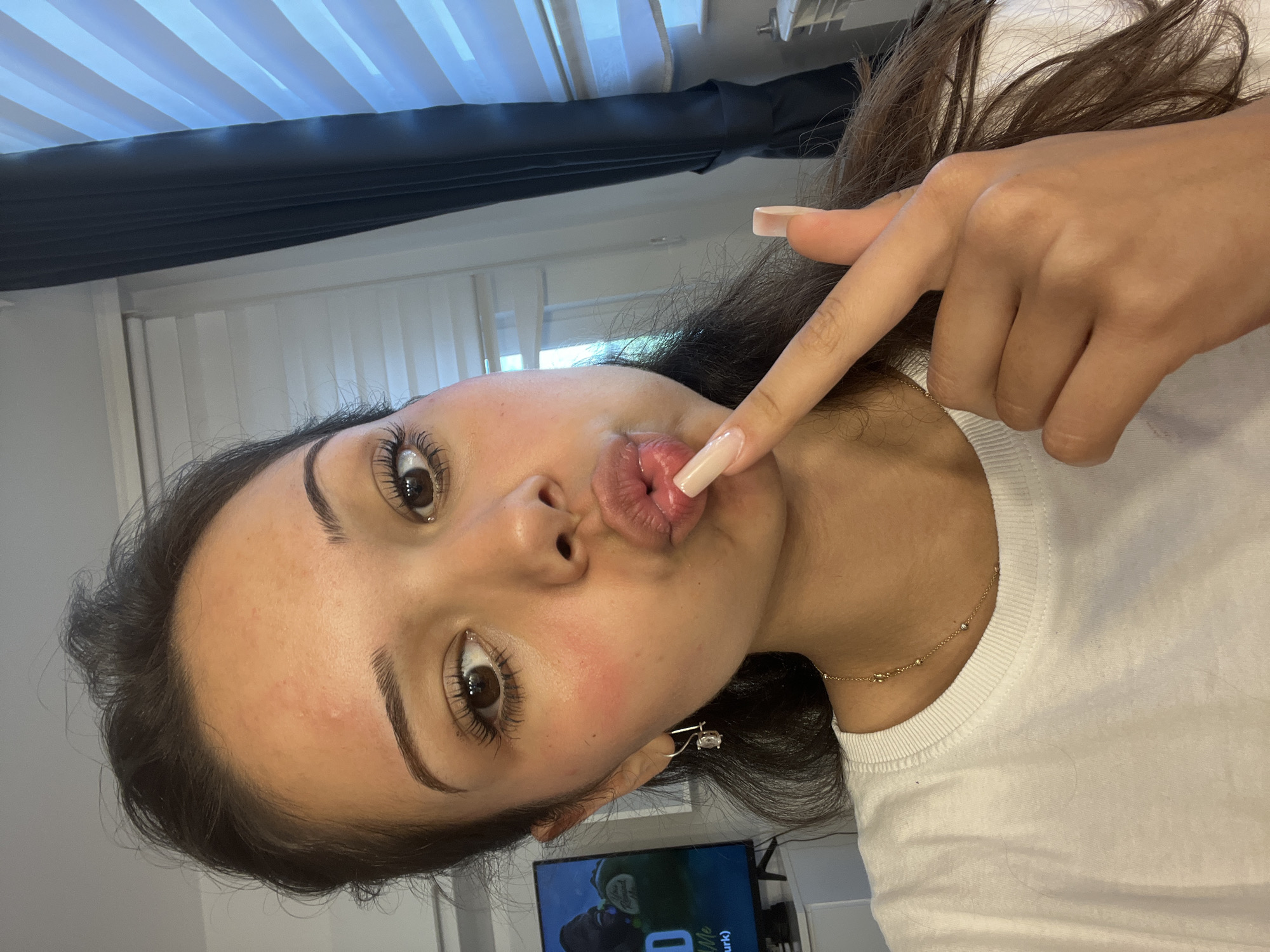 A young woman with brown hair and brown eyes is making a kissing face towards the camera. She is wearing a white t-shirt and a gold necklace. She has on long, light-colored fingernails, and is touching her face with her index finger. Her left cheek has a blush of pink. There is a small, clear earring in her left earlobe. In the background, a television screen displays an image of a man. A window with horizontal blinds is visible to the right. The woman's pose suggests a selfie taken indoors. This image was captured in Zlatograd, Bulgaria.