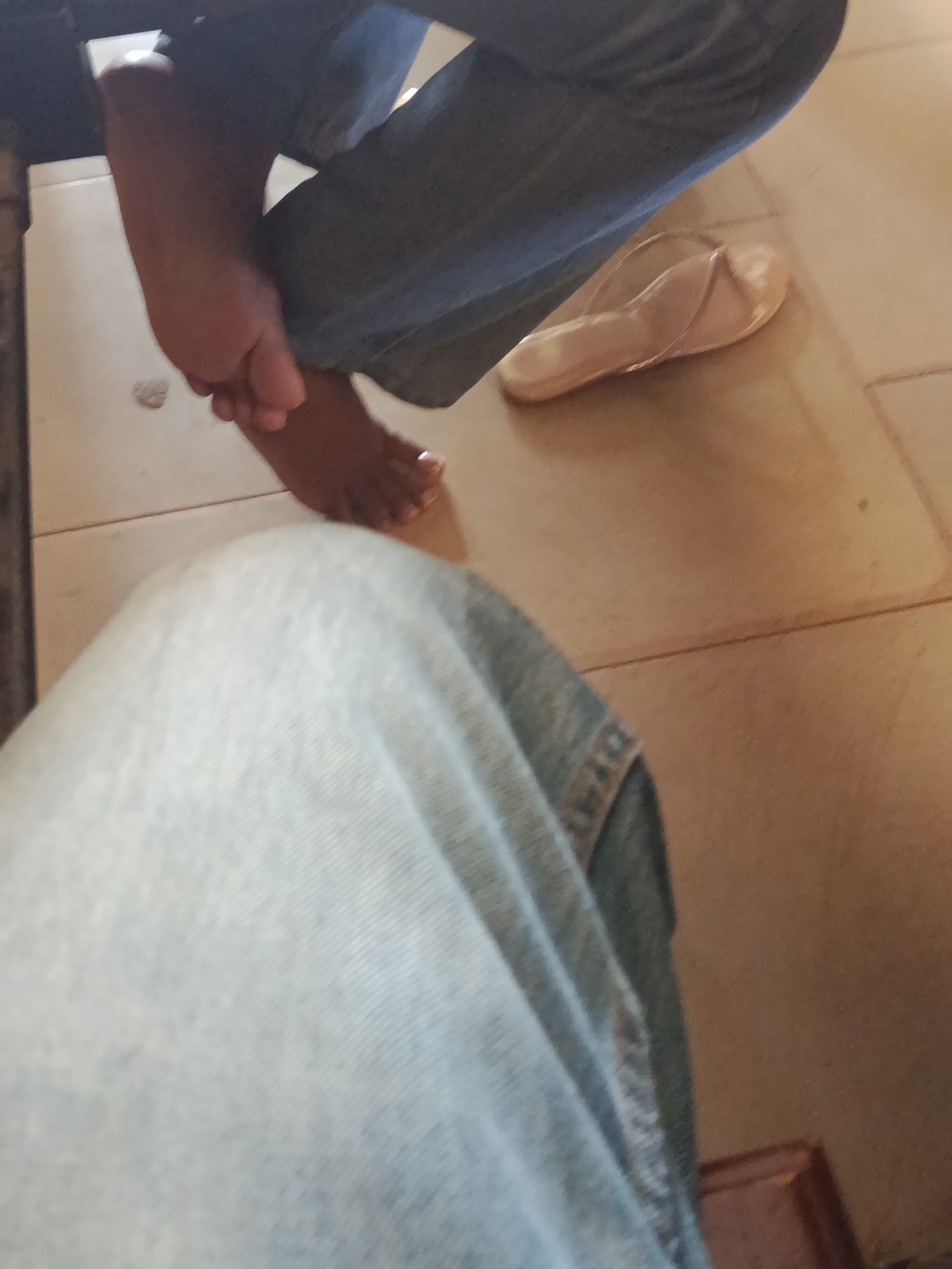 In this close-up, low-angle shot, two individuals, both wearing denim jeans, are seated with their legs crossed. One individual's bare feet are visible, with the soles of their feet turned upward. Their toes are clearly defined, and the skin appears to be a medium brown tone. The other individual's foot is clad in a light beige flip-flop. The flip-flop has a thin strap that goes between the toes and across the top of the foot. Both individuals are seated on a floor tiled with square, light brown tiles. The lighting in the image is dim, creating a muted color palette. The overall scene is unremarkable and appears to be an indoor setting.