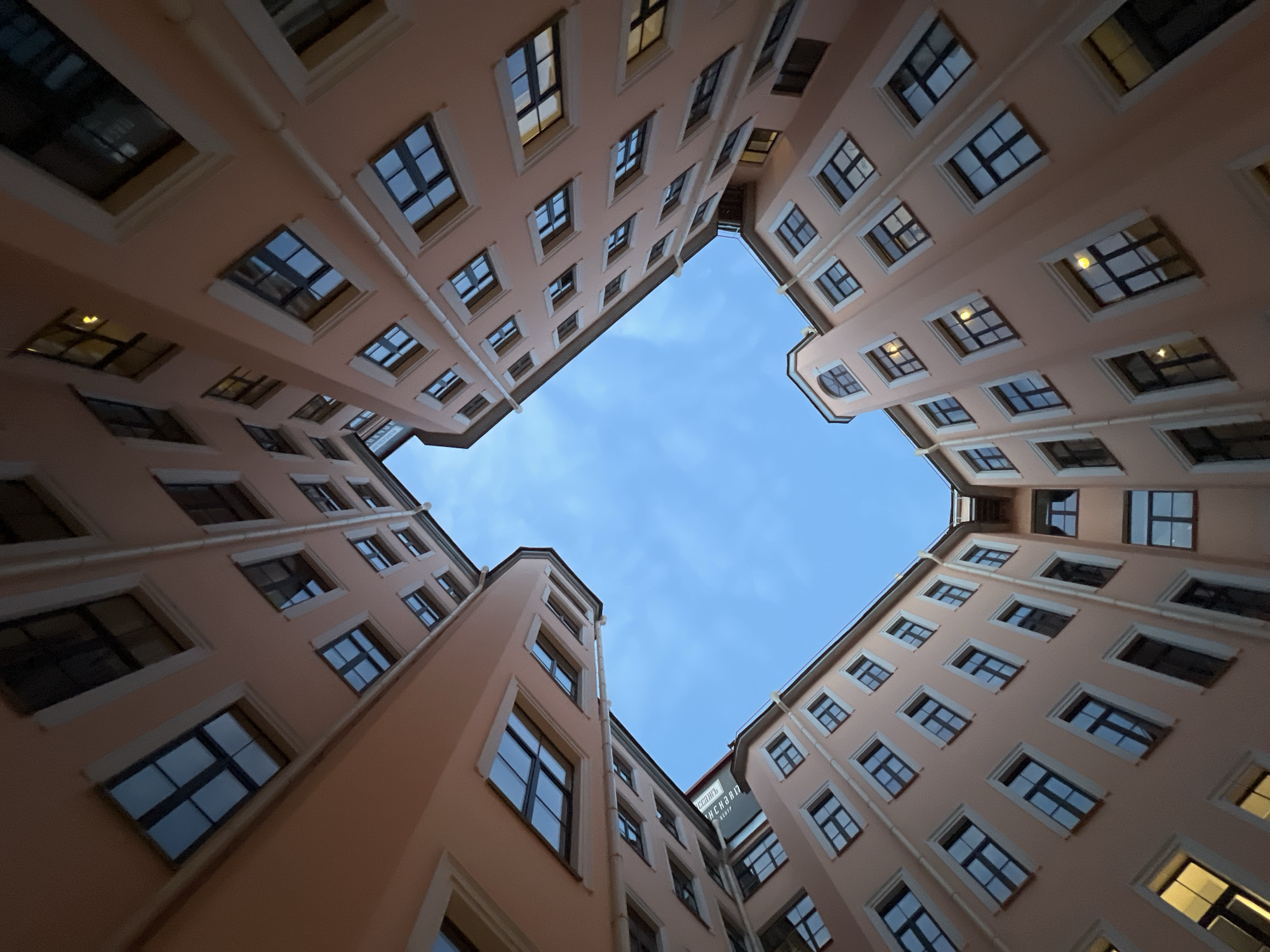 The image depicts a courtyard in Saint Petersburg, Russia, enclosed by buildings with peach-colored facades. The perspective is from below, looking up towards the sky, which appears blue with thin wisps of clouds. The buildings are rectangular, multi-storied structures, with evenly spaced windows, most of which are rectangular with black frames. Some windows reflect light, indicating they might be illuminated from within. Vertical drainpipes run down the exterior walls of the buildings. The buildings form a square shape, framing the sky above. A sign is visible on one building.