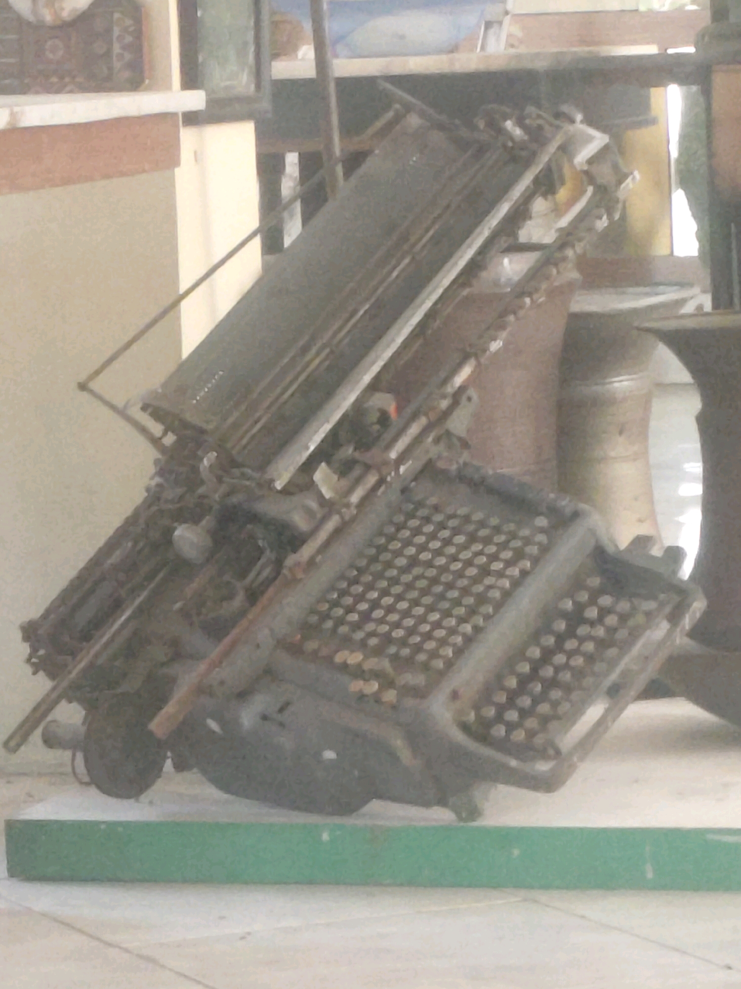 The image depicts a close-up, angled view of a heavily rusted and partially disassembled vintage typewriter, possibly a Braille typewriter given the array of raised dots on the keyboard. The typewriter is positioned outdoors on a tiled surface, with a green reflective object, possibly a bench or a decorative element, in the foreground. In the background, a cream-colored wall is visible, with architectural details that suggest it could be part of a building. The overall impression is one of decay and abandonment, with the object likely an artifact of historical or industrial significance, possibly located in an area subject to the elements. The context of Cisaga, Indonesia, implies a tropical or subtropical climate, which could contribute to the corrosion and wear observed on the typewriter. No individuals are present in the image.