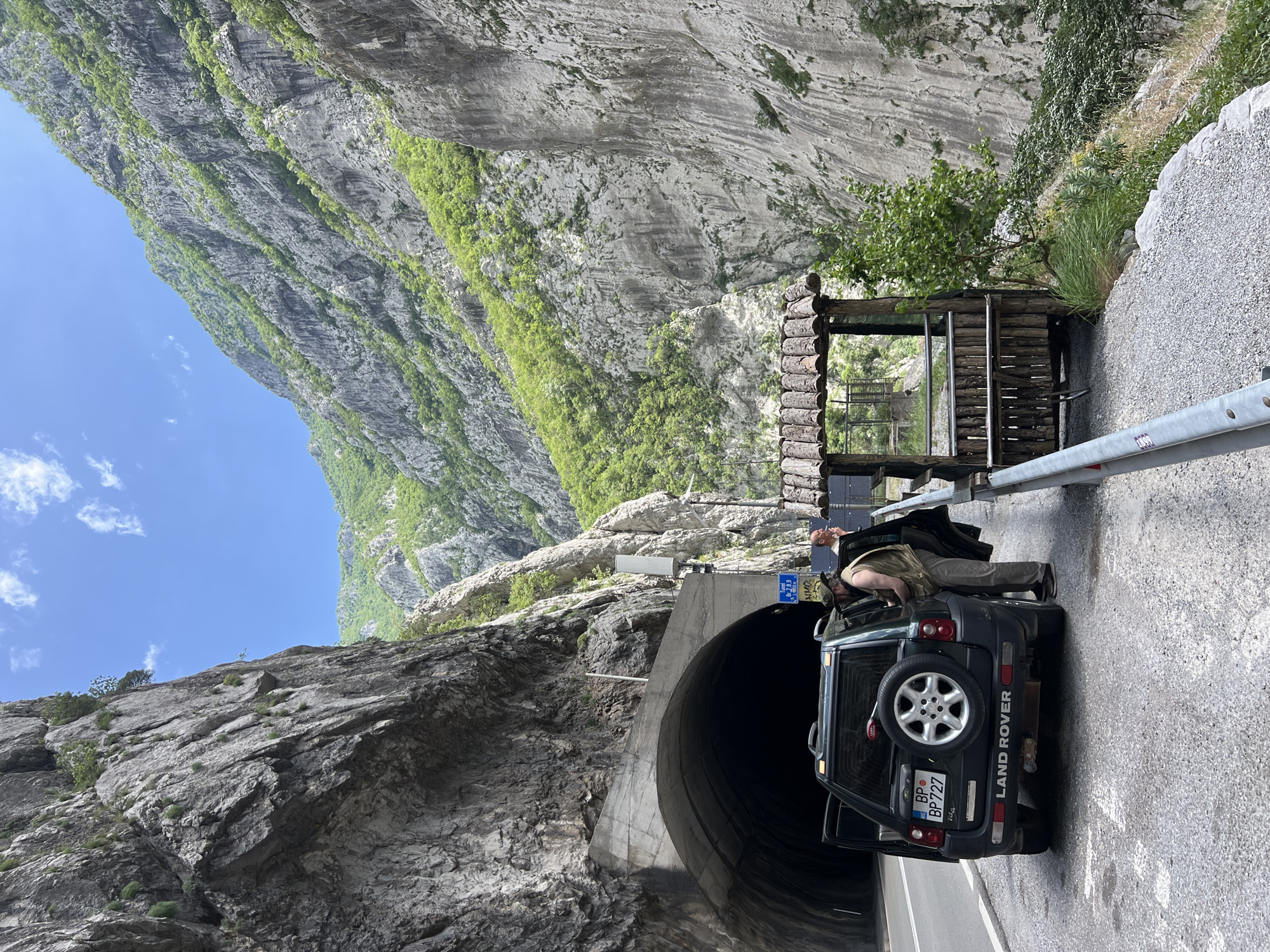 The image depicts a mountainous landscape with a road and a tunnel entrance. A dark-colored Land Rover vehicle is parked next to the tunnel entrance, with its rear door open. A person is leaning into the vehicle, appearing to be interacting with something inside. Adjacent to the road is a wooden structure resembling a small shelter or viewing point. The scene is likely located near Vlahovići, Montenegro, judging by geographical cues.