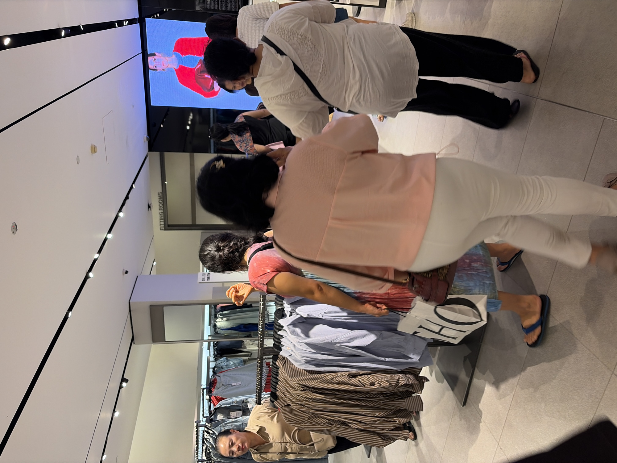In Kuta, Indonesia, within a retail establishment, several individuals are observed in a well-lit store environment. The scene appears to be a clothing store, evidenced by racks of garments displayed. In the foreground, a person wearing a pale pink top and white pants is walking away from the camera. To their left, another individual in a white hooded top and black pants is also walking. A large screen displays a fashion image, featuring a woman in a red jacket.
Further into the store, a woman in a beige shirt and a crossbody bag is standing near a clothing rack displaying horizontally striped and vertically striped shirts, as well as light blue shirts. To her right, a woman wearing a pink and tie-dye top and a skirt is holding a white shopping bag with black lettering that reads "HO." She is also wearing blue flip-flops.
The environment is characterized by tiled flooring and ceiling-mounted track lighting. A sign indicating "FITTING ROOMS" is visible in the background. The overall impression is of a typical shopping scene in a retail setting.