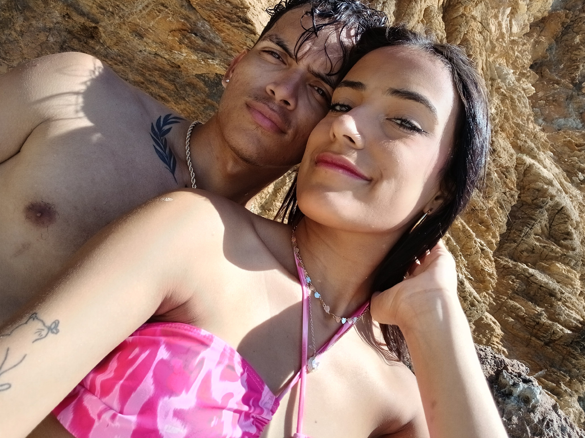 A close-up, sunlit selfie captures a young man and woman posing together against a backdrop of textured, brown rock formations. The setting appears to be an outdoor, natural environment, possibly a rocky coastline or beach in Altagracia, Venezuela, under clear, sunny conditions with strong daylight casting visible shadows.
The man is positioned behind the woman, his head resting near hers. He has dark, wet, curly hair, a light mustache, and is shirtless, displaying a silver chain around his neck and a distinct tattoo of a leafy branch on his left shoulder/collarbone area. He looks directly at the camera with a calm, slightly smiling expression.
The woman is in the foreground, closer to the camera, partially obscuring the man. She has long, dark hair, some of which she holds with her left hand, and is wearing a vibrant pink patterned bikini top. Two delicate necklaces adorn her neck: one with small heart charms and another with a larger, sparkling pendant. Small, faint tattoos, including a feline-like figure, are visible on her left arm. She is looking towards the camera with a gentle smile and a relaxed demeanor. Both individuals exhibit contented expressions, suggesting a pleasant moment captured.