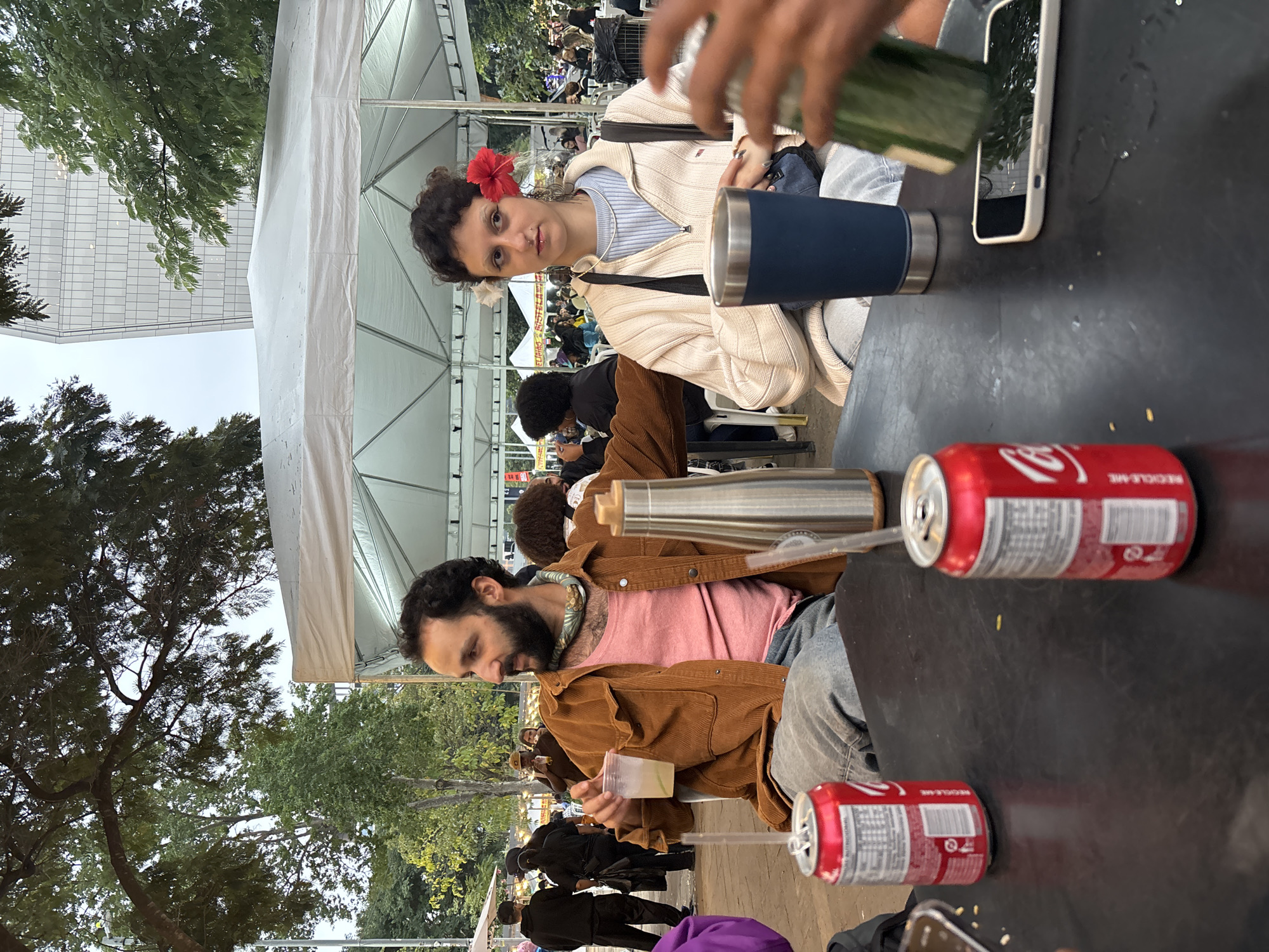 A medium-shot captures two individuals seated at a black table under a tent, likely at an outdoor event. A woman with dark curly hair and a red flower in her hair wears a light-colored zip-up sweater and gazes at the camera. A man with a beard, wearing a pink shirt and a brown corduroy jacket, looks down at a translucent plastic cup in his hands.
On the table are two red cans of Coca-Cola, a stainless steel water bottle, and a dark blue Yeti cup. A white smartphone with a case also rests on the table. In the background, other people are visible seated at tables, suggesting a populated event.
Trees are visible in the background, as well as a tall building. The location is identifiable as São Paulo, Brazil.