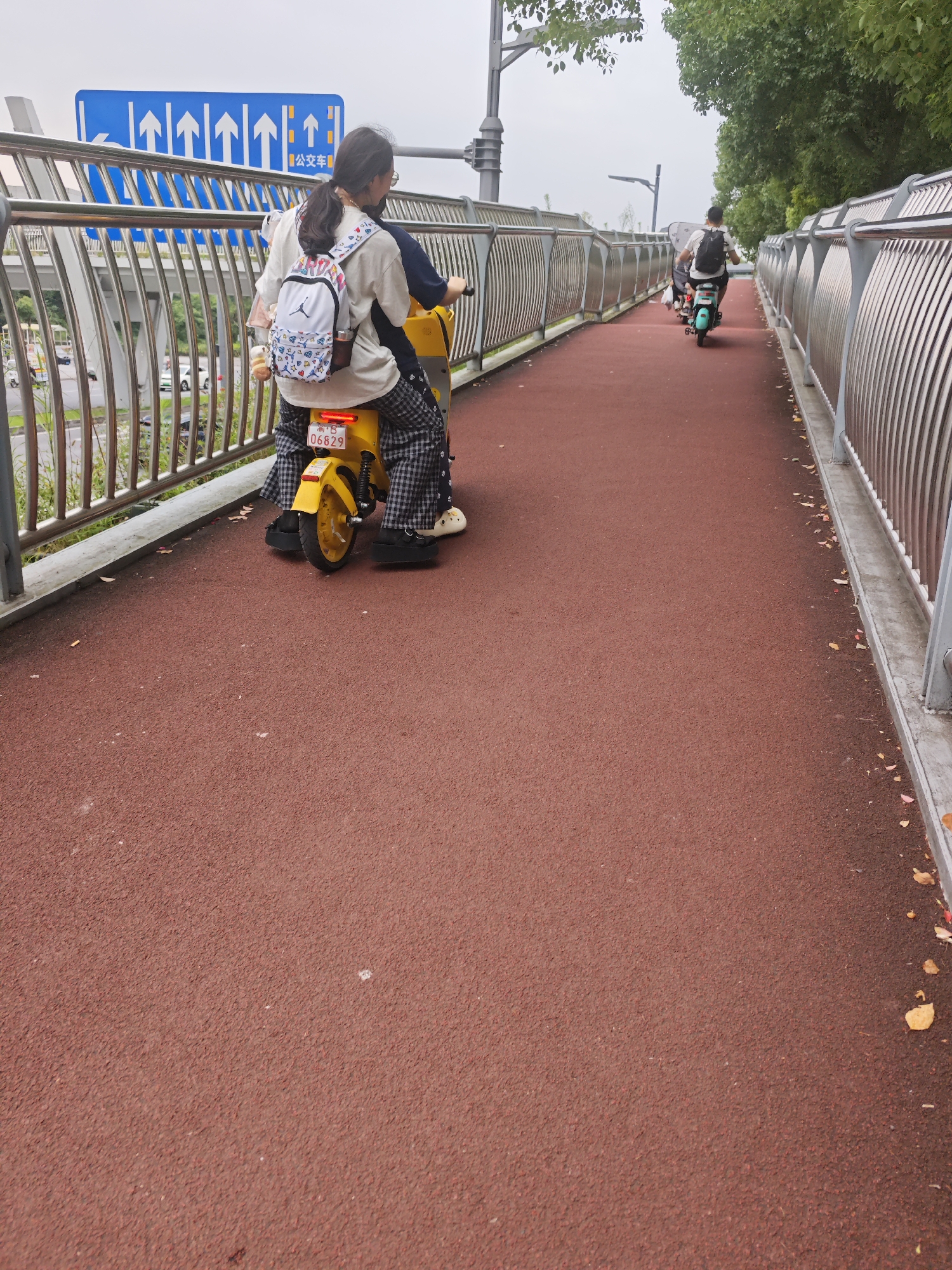 Two people, a male and a female, are riding on a yellow electric scooter on a red asphalt path, presumably a pedestrian or bicycle path, which is bordered by a metal railing. The female is seated in front, wearing a white t-shirt, checkered pants, and a backpack. The male is behind her, wearing a dark shirt and a backpack. In the background, there is traffic on a road below the elevated path, with cars and trucks visible. A blue traffic sign with multiple upward-pointing arrows and Chinese characters is prominently displayed on the left side of the path. The sky is overcast, suggesting it might be a cloudy day. The location is identified as the city of Chongqing High-tech Zone, China.