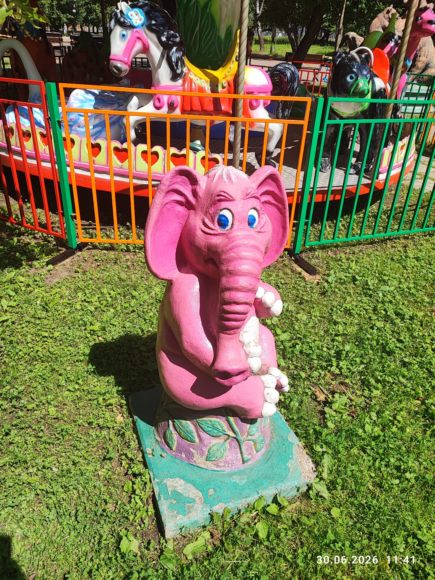 A pink elephant statue sits in a grassy area, its blue eyes wide and its trunk curled. It is positioned in front of a brightly colored carousel, which is partially visible. The carousel features a horse with a black mane and pink halter, a black bear-like creature with green accents, and other animal figures. A colorful fence, with vertical bars of green, orange, and red, separates the carousel from the foreground. The grass is green and appears well-maintained, with some patches of clover. Sunlight casts shadows on the ground, suggesting it is daytime. The scene appears to be an outdoor amusement park or a public recreation area. The date "30.06.2026 11:41" is visible in the lower right corner of the image, indicating a timestamp.