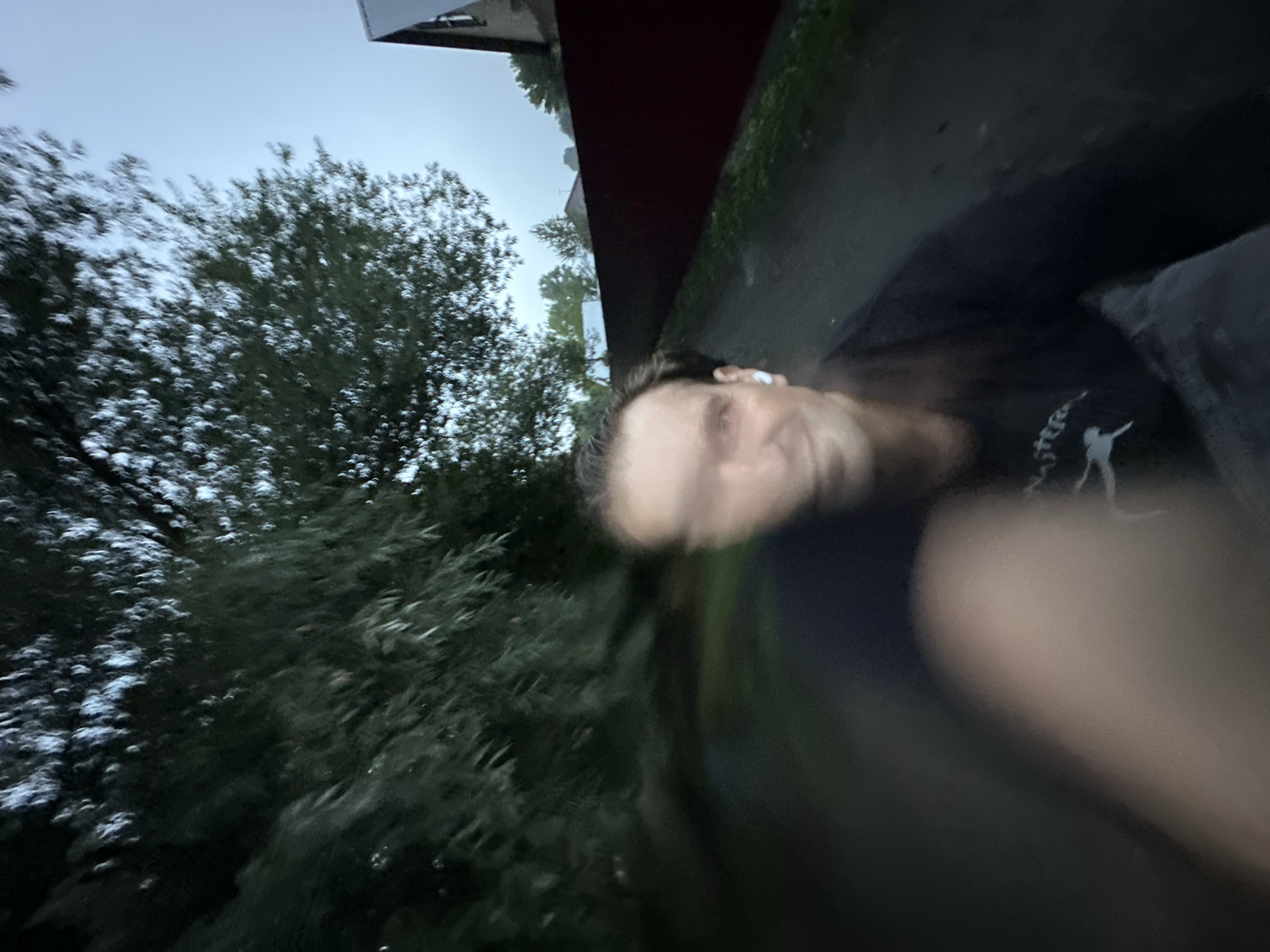 The image depicts a person with their face blurred due to motion, wearing a dark long-sleeved shirt and what appear to be white wireless earbuds. The background suggests an outdoor environment, possibly a rural or suburban area. Trees with green foliage are visible, along with a dark red fence and a portion of a building with a triangular roof. The ground appears to be a dirt or gravel path. The sky is overcast and the lighting suggests it is either dawn or dusk. The overall impression is of someone in motion, perhaps cycling or running, capturing a selfie. The location is identified as Biysk, Russia.