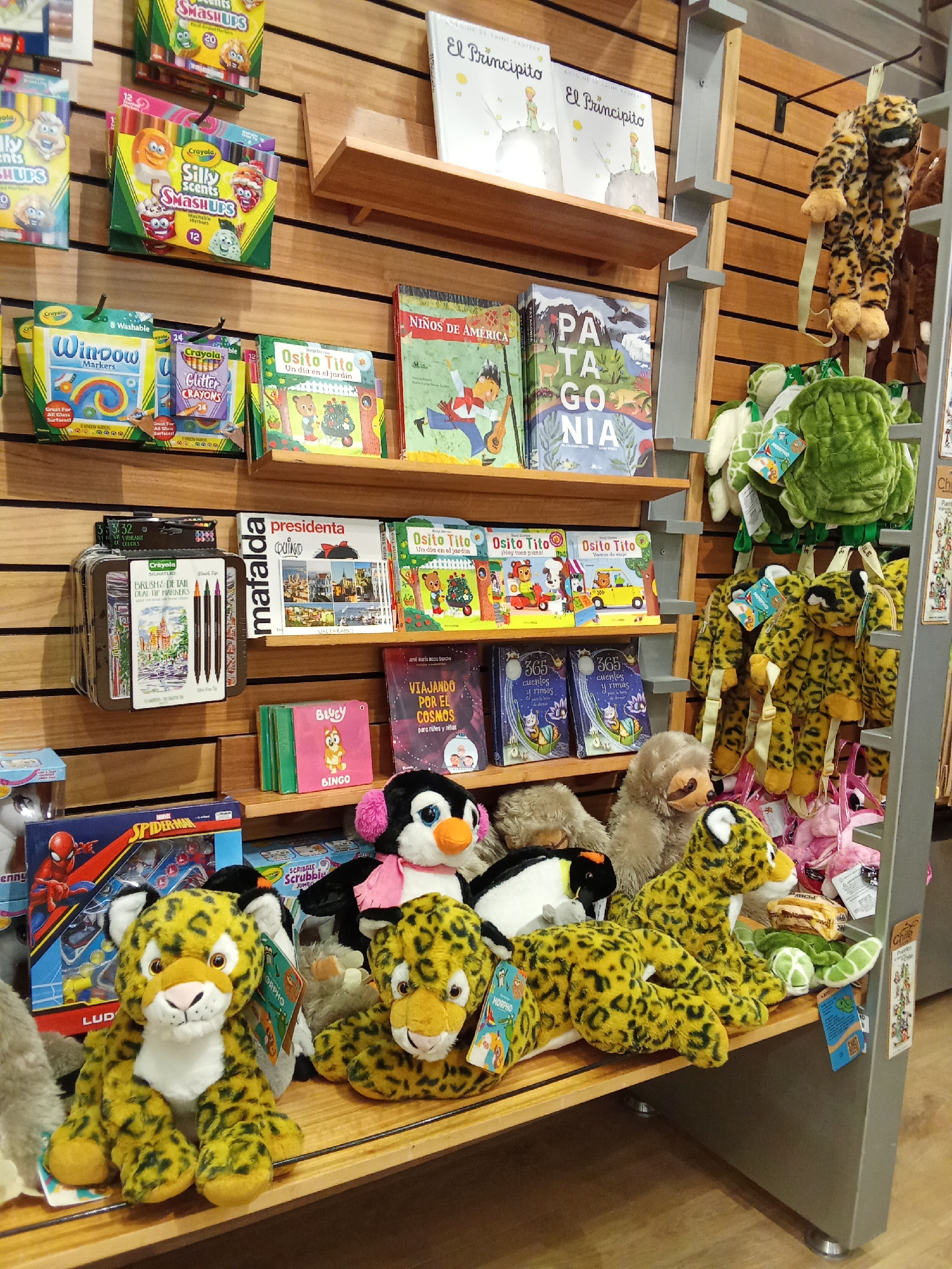 The image depicts a retail display of children's books and plush toys, likely in a store. The display features wooden shelving filled with a variety of items. On the top shelf, several books are visible, including "El Principito" by Antoine de Saint-Exupéry. The middle shelves showcase more books, such as "Osito Tito" and "Patagonia", alongside art supplies like markers and crayons. Plush toys, predominantly animal-themed, are arranged on the lower shelves and the floor. Among them are stuffed jaguars, a penguin, a sloth, and turtle-shaped pillows. The background shows a wooden wall paneling, typical of a retail environment. The scene is brightly lit, suggesting an indoor setting. There are no people visible in the image. Based on the context provided in the problem description, this retail display is located in the city of Pudahuel, Chile.