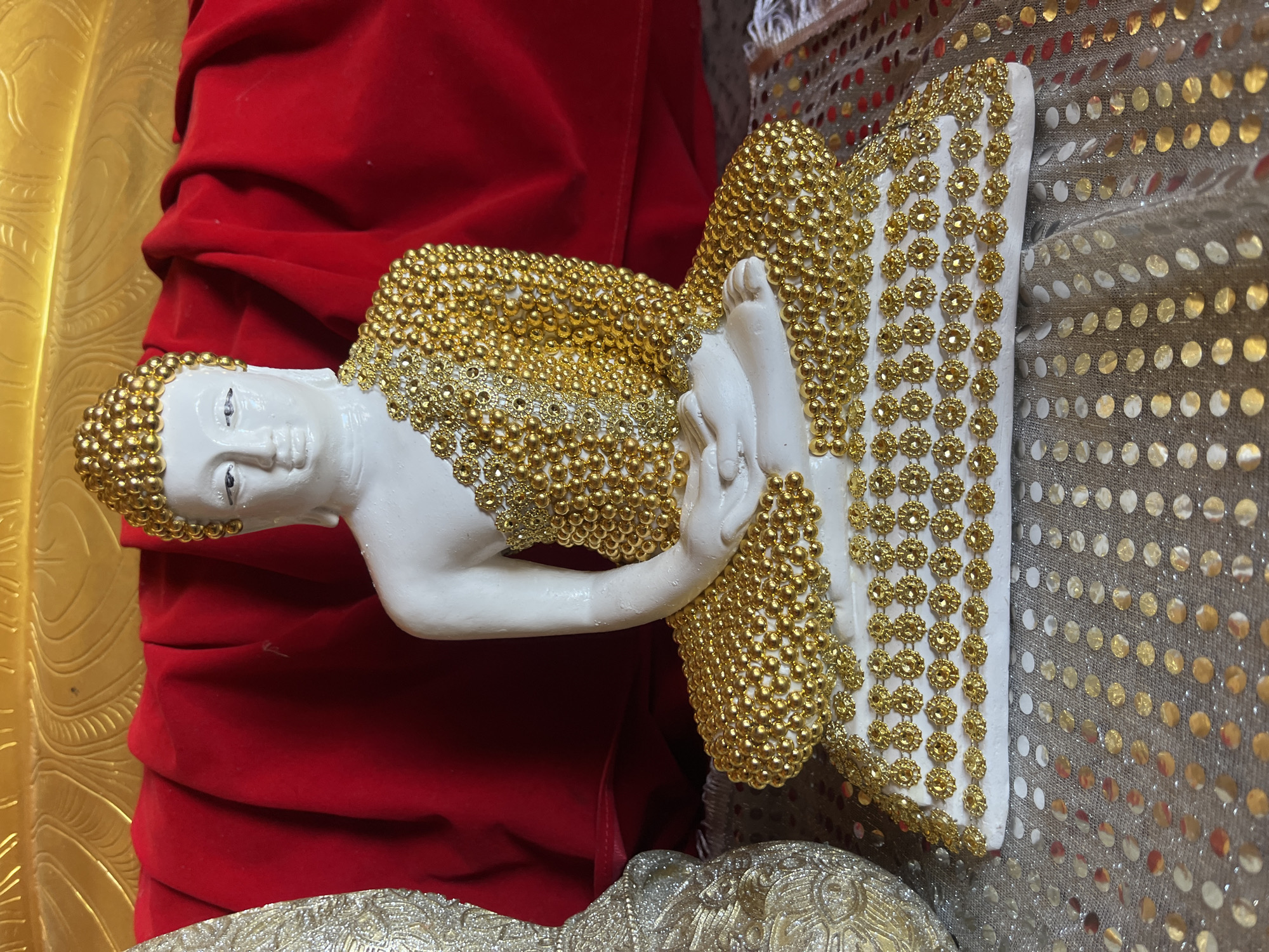 A white statue of Buddha is seated in a meditative pose, adorned with numerous gold beads. The statue is positioned in front of a red draped fabric. To the left of the Buddha statue, a portion of a decorative, ornate object covered in gold and silver embellishments is visible. The foreground consists of a textured surface with scattered circular metallic sequins in gold and silver. The background features a large, gold-colored structure with textured patterns. The scene appears to be an indoor setting, possibly a place of worship or a decorated display, with no people present. The overall impression is one of opulence and religious reverence.