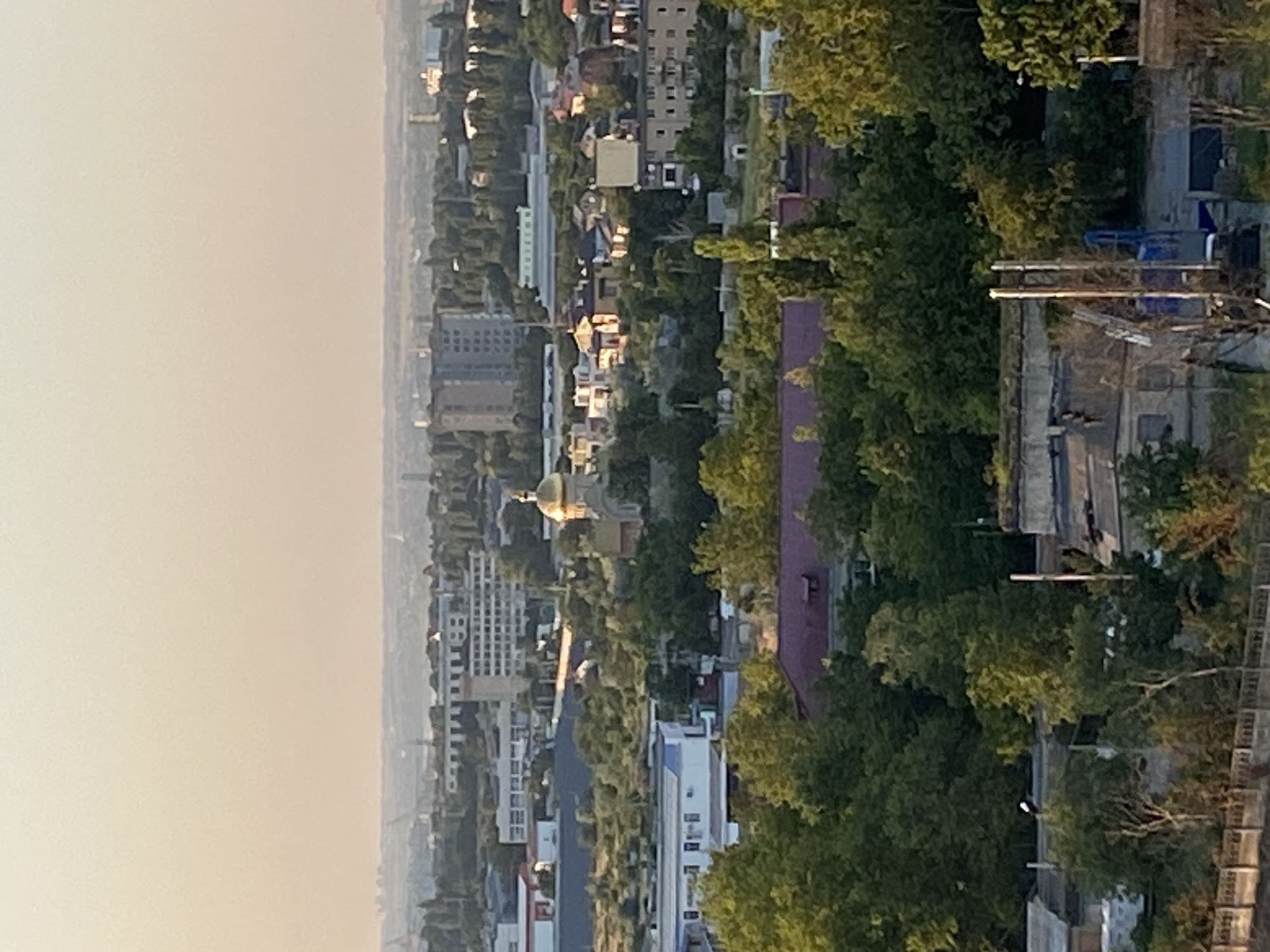 The image is an aerial view of a city with a hazy sky. In the foreground, there are dense green trees. Beyond the trees, there are various buildings, including residential and industrial structures. A prominent golden-domed building, likely a church, is visible in the mid-ground. In the background, more buildings and infrastructure stretch towards the horizon. The scene appears to be in a suburban or industrial area of Chornomorsk, Ukraine. There are no people visible in the image. The overall atmosphere suggests an overcast day with low visibility.