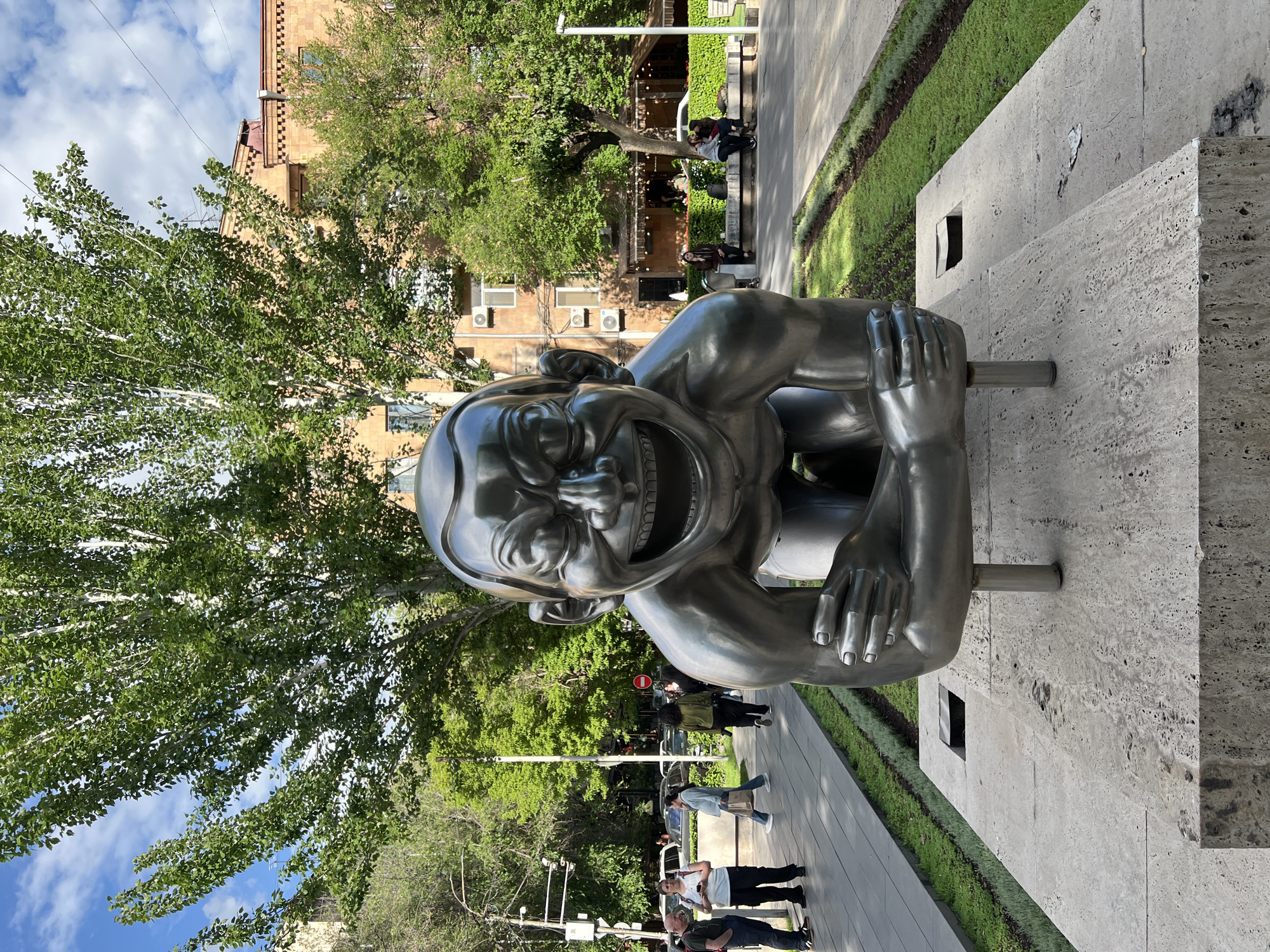 The image shows the "Men" monument in Yerevan, Armenia. This statue is a large, grinning, and crouching figure. It is made of metal and is placed on a stone platform along a pedestrian walkway. Trees surround the area, with buildings visible in the background. People are walking and standing nearby. The sky is partly cloudy.