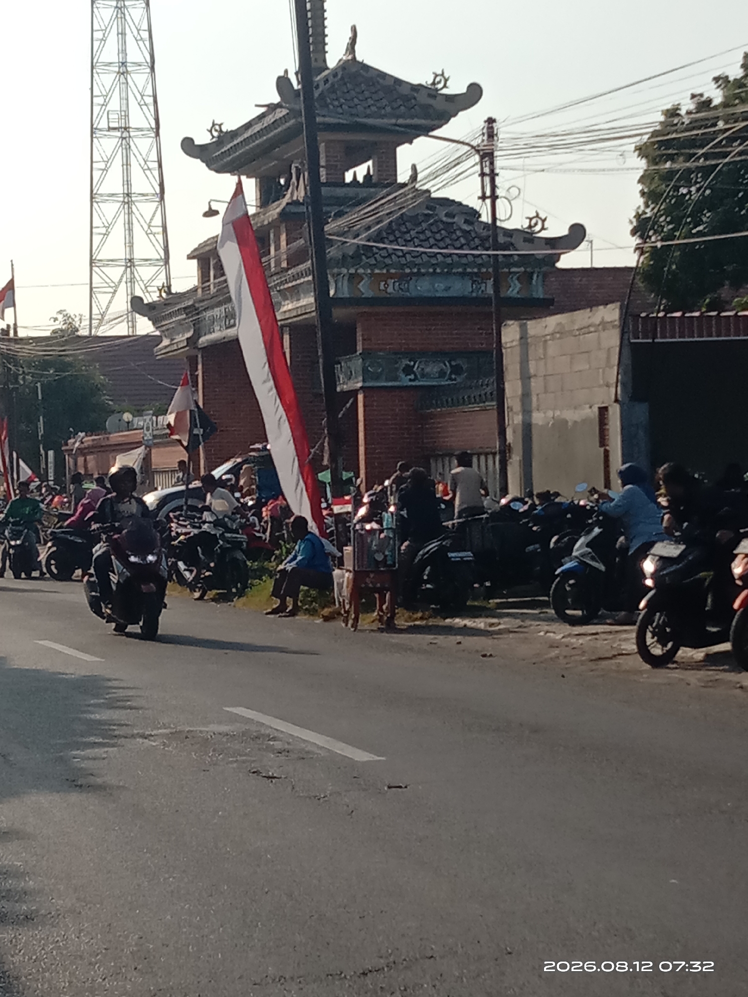 The image depicts a street scene in Gudo, Indonesia, on August 12, 2026, at 07:32. The foreground shows a paved road with white lane markings, some of which are faded, and visible potholes. In the background, a brick structure resembling a temple gate or traditional Indonesian building with a tiled roof is prominent, adorned with two large Indonesian flags. To the left of the gate, a tall radio tower is visible against a bright sky. A crowd of people are gathered around numerous parked motorcycles on the side of the road, with some individuals riding or about to ride their vehicles. The atmosphere appears to be one of activity and perhaps an event or gathering.