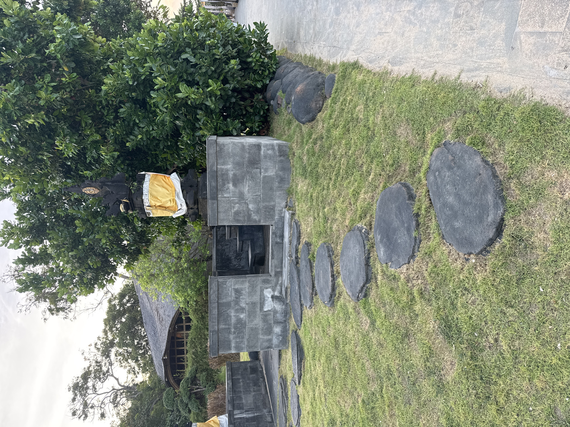 The image captures a scene of a garden path in an unknown location. A stone pathway is lined with round, dark stepping stones embedded in the green grass. Adjacent to the path, a gray, multi-tiered stone structure stands. Yellow and white fabric is draped over part of the structure. Lush green vegetation surrounds the stone structure, and a building with a curved roof is visible in the background. A paved walkway borders the grassy area. The sky is overcast.