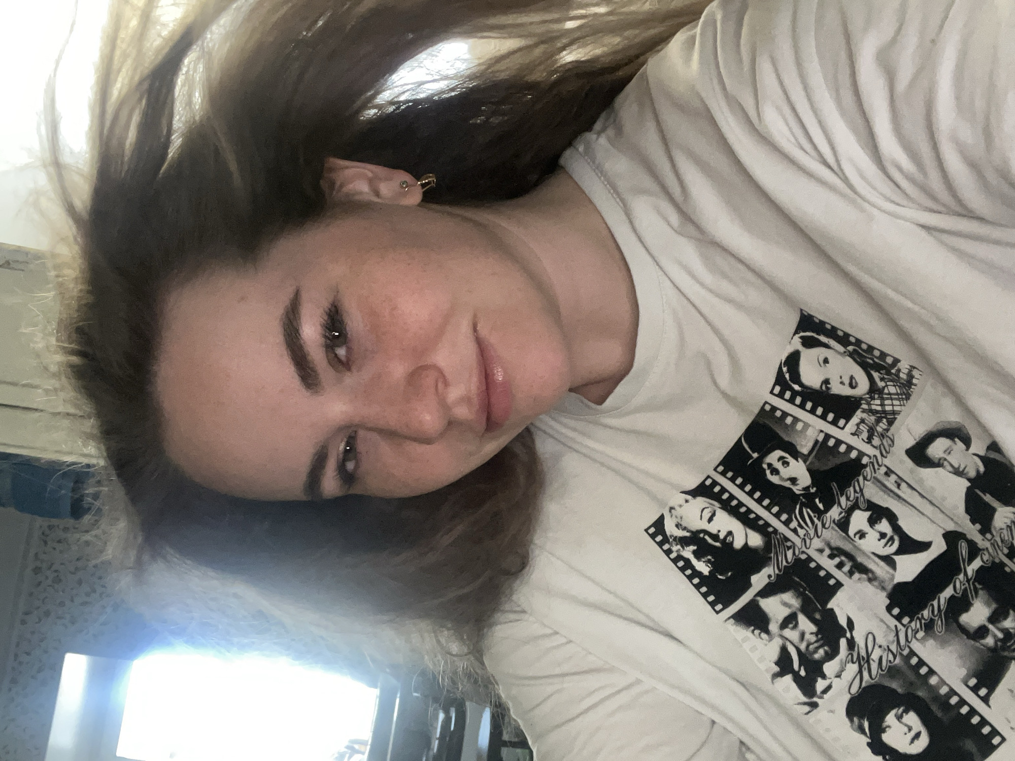 A selfie depicts a woman with brown hair and fair skin wearing a white t-shirt with black and white images of movie legends in a filmstrip design. The camera angle is tilted, and the background is blurry, suggesting an indoor setting, possibly in the city of Chaikovsky, Russia.