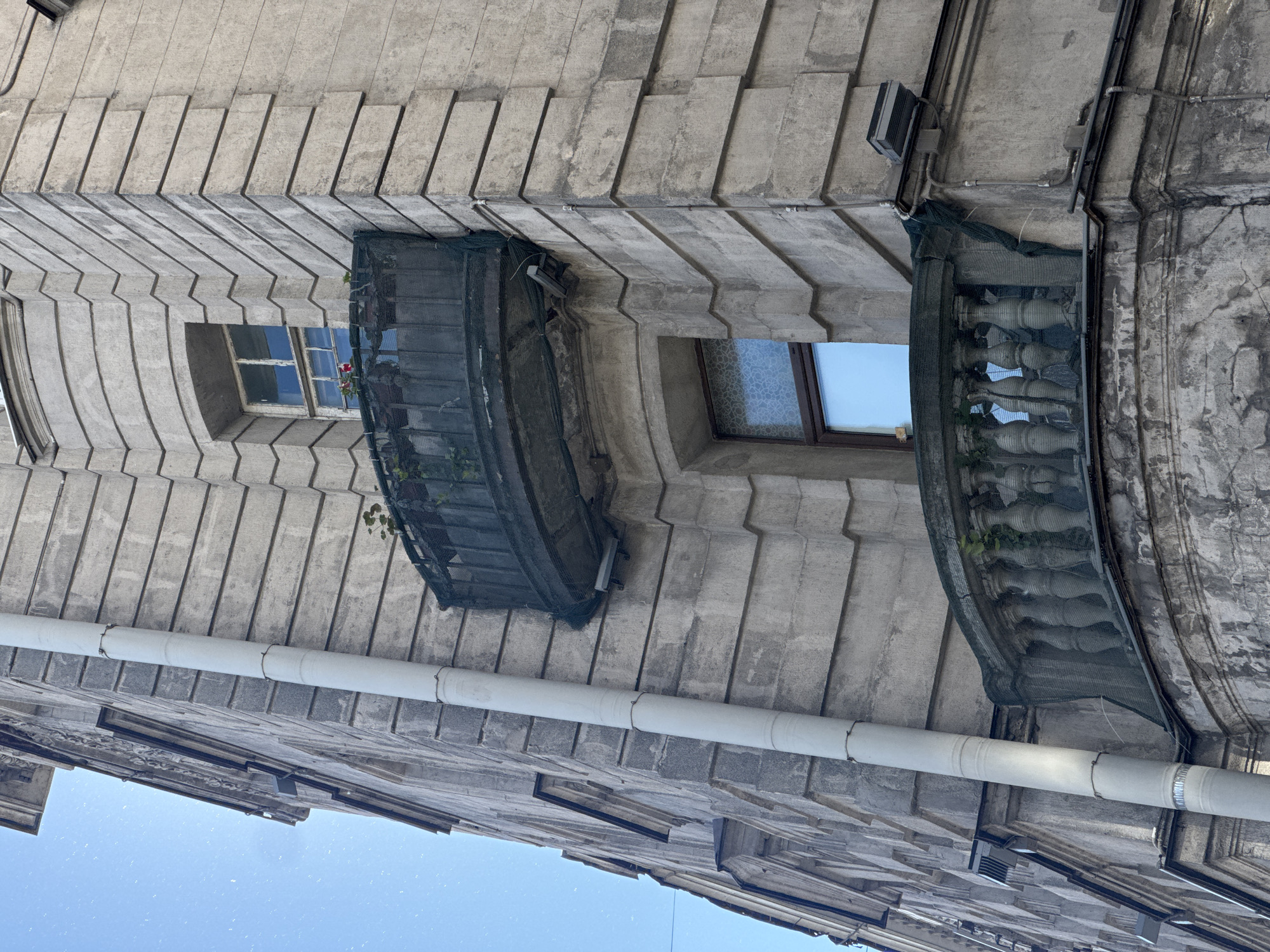 The image captures a close-up, low-angle view of the exterior of an old, stone building on a clear day. The building features ornate architectural details, including horizontal stone banding and decorative moldings. Two balconies are visible: an upper, curved balcony with what appears to be a dark mesh covering, and a lower, semi-circular balcony with classical balusters. A large, white downspout runs vertically along the left side of the building. The sky, a clear blue, is visible in the upper left quadrant of the image. The stone facade shows signs of age and weathering.