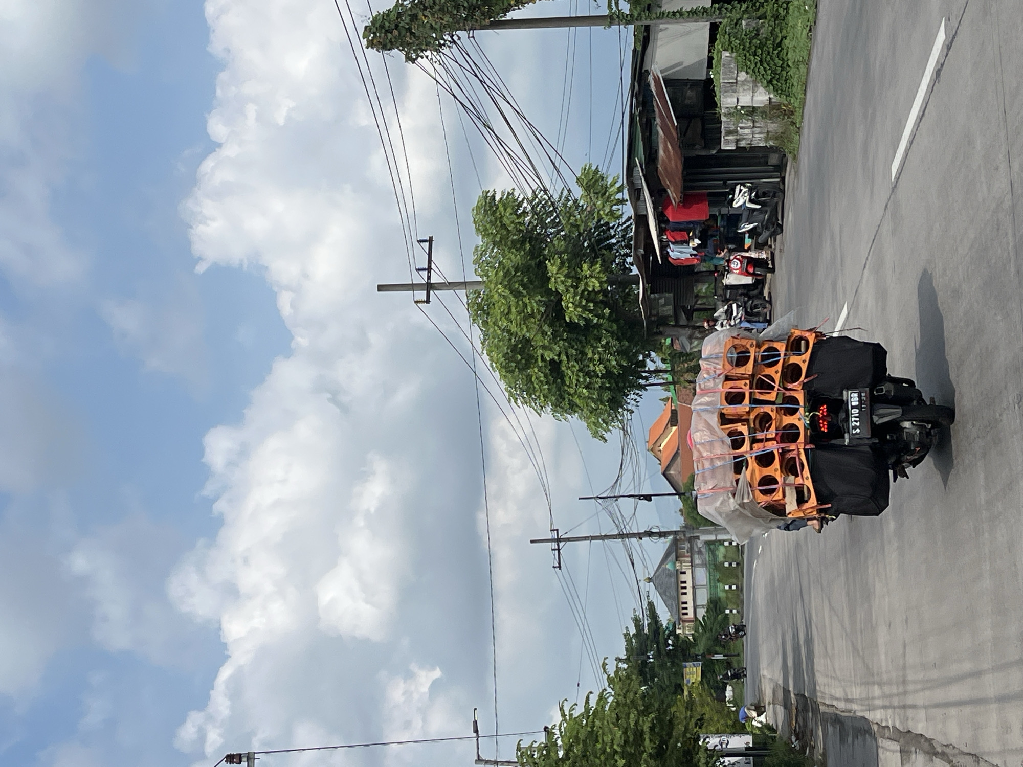 A motorcycle laden with a large, orange cargo covered in plastic sheeting travels down a paved road in Kepatihan, Indonesia. The motorcycle is viewed from behind, and the cargo appears to be stacked in multiple layers. The rider, wearing a helmet, is partially obscured. The road is lined with trees and buildings, including a structure with a domed roof in the distance. Overhead, power lines crisscross the sky, which is filled with scattered white clouds. Several other motorcycles and people are visible in the background near a small shop selling clothing.