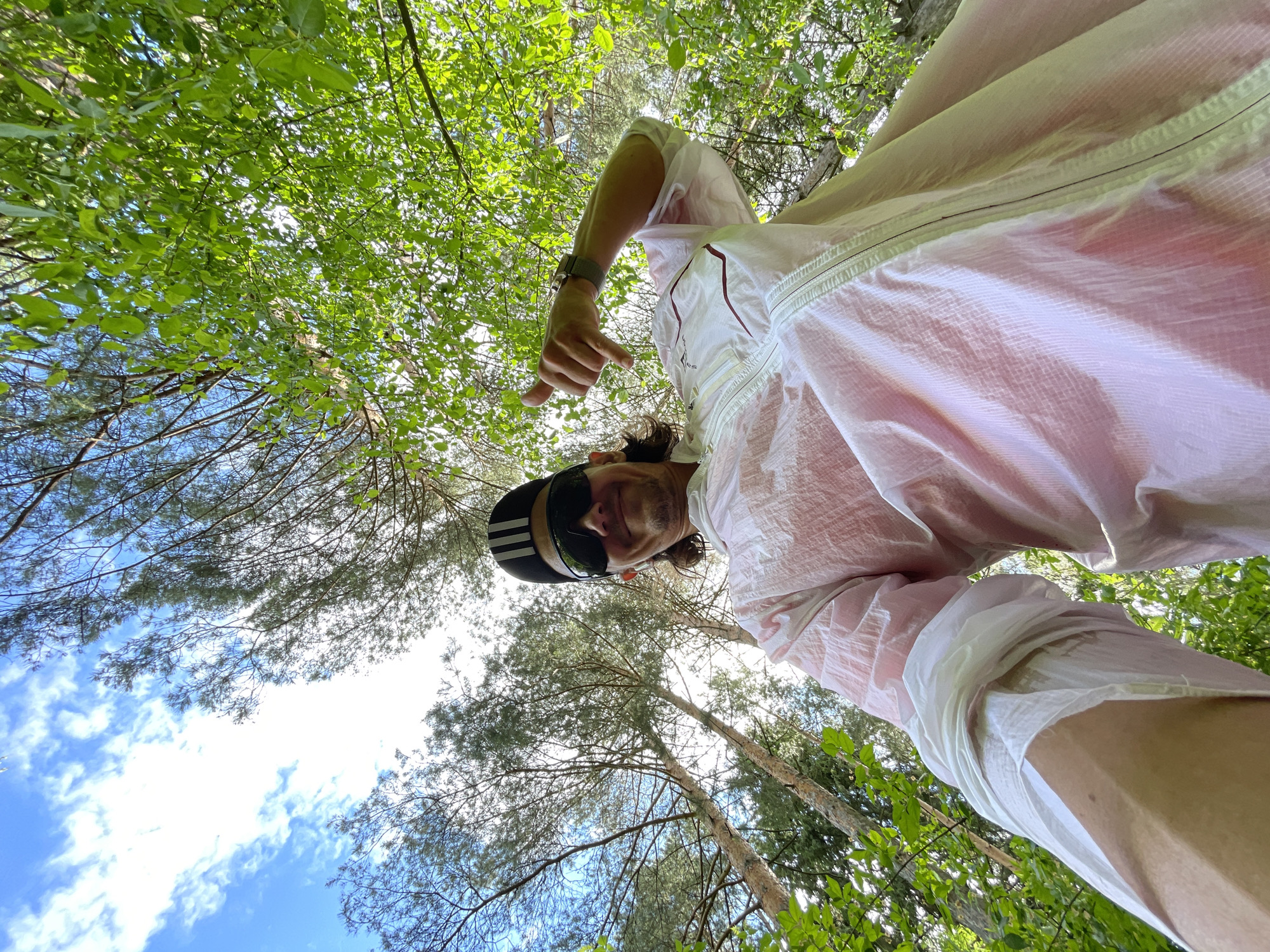 A low-angle shot captures a person in a white and pink shirt, wearing sunglasses and a black and white striped hat. The person points downward with their right hand. The backdrop features a canopy of green trees, with glimpses of a bright, partly cloudy sky visible between the branches. The location appears to be a wooded area, likely a park or forest, in the city of Unknown, Unknown.