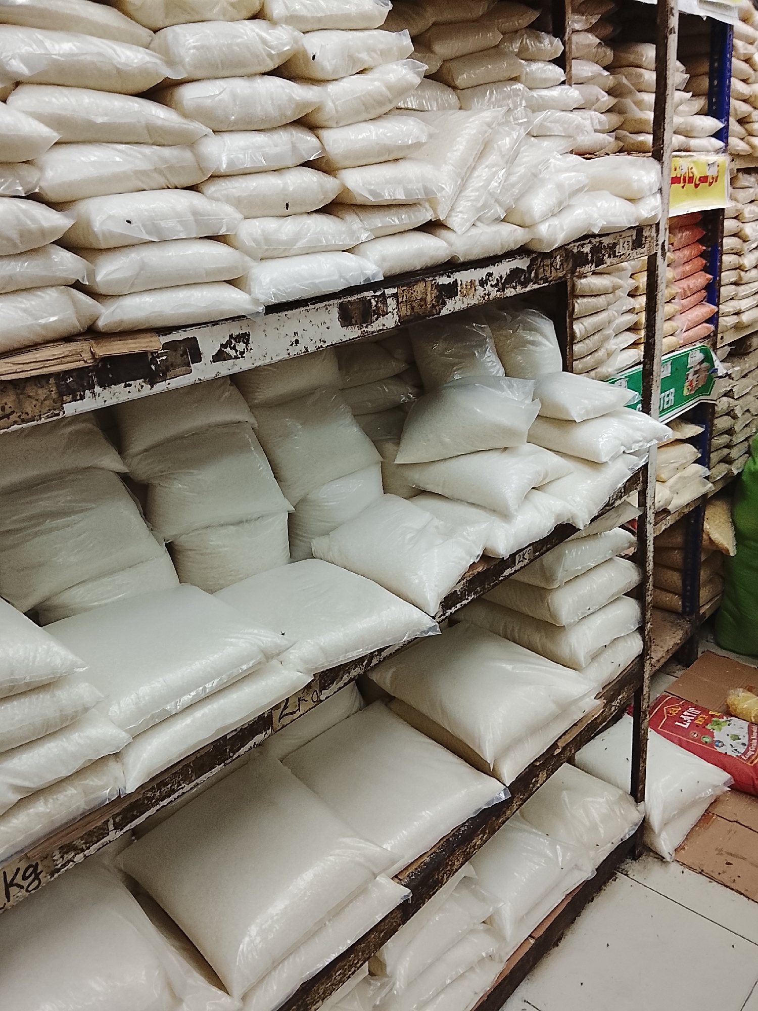 The image depicts a grocery store shelf in Gulgasht Colony, Pakistan. The shelves are stacked with multiple white plastic bags, likely containing sugar or rice. The shelves are constructed from dark metal. The bags appear to be uniformly shaped and arranged in stacks. Other grocery items are visible in the background, including stacks of lentils and rice bags. A red bag of “LATIF RICE” sits on the ground near the bottom shelf. The shelves are old and rusty. The floor is light-colored tile.