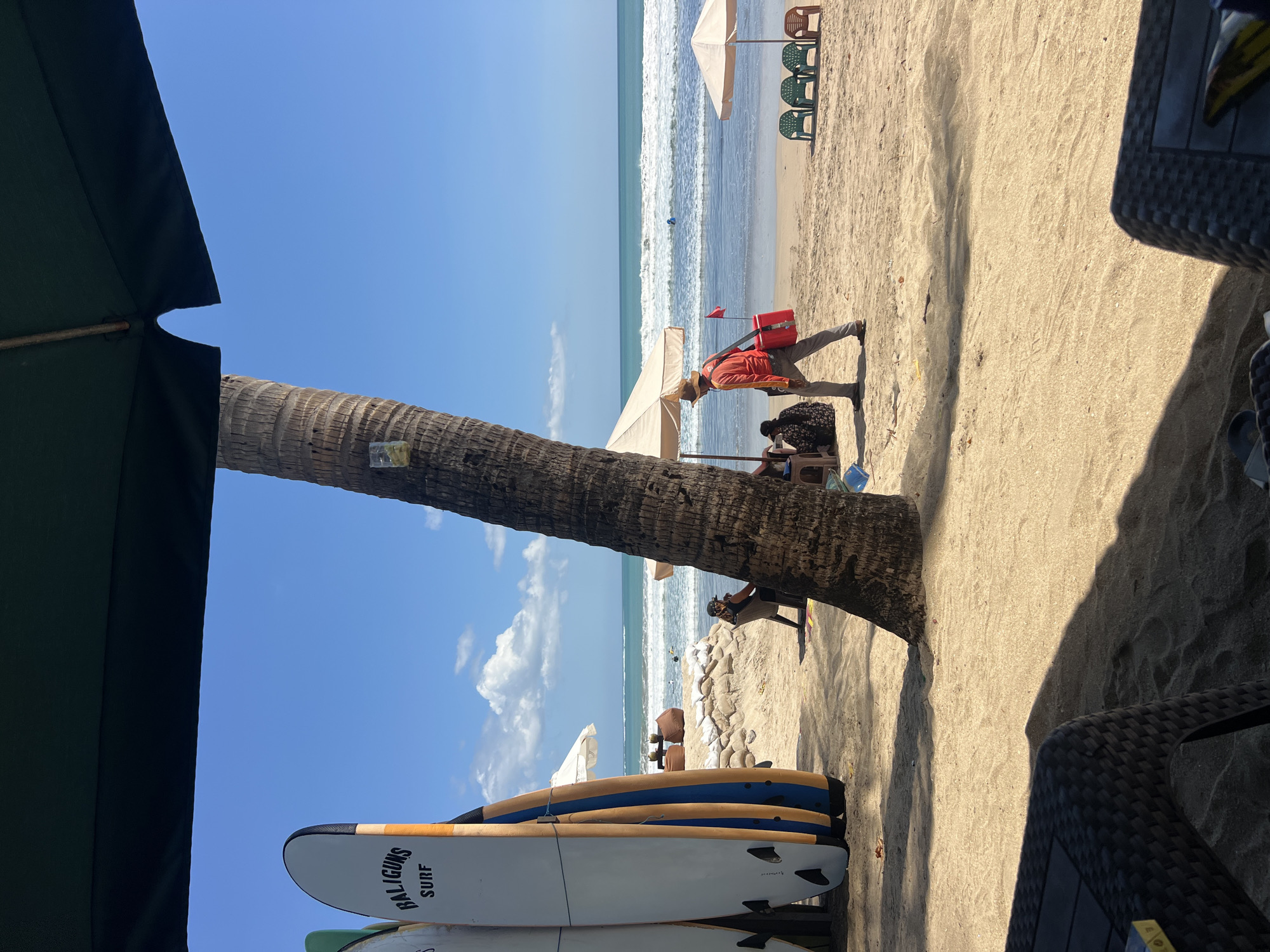 This image depicts a scene on Kuta Beach, Bali, Indonesia, on a sunny day. The foreground is dominated by the shadowy underside of a large umbrella and a portion of a dark, woven table, suggesting a seating area at a beachside establishment.
In the mid-ground, a sandy beach stretches towards the ocean. Several surfboards, two of which are labeled "BALIGUNS SURF," stand upright, leaning against each other. Further back, two people are seated on the sand under a beige umbrella. A man, wearing a wide-brimmed hat and an orange shirt, walks across the sand carrying a red cooler bag over his shoulder. He is walking away from the viewer and towards the ocean. In the distance, the turquoise ocean meets a bright blue sky with scattered white clouds. Waves are rolling onto the shore. Several colorful plastic chairs are set up on the sand near the water's edge, some under small, beige umbrellas. The presence of surfboards, beach umbrellas, and chairs indicates a typical tourist beach setting in Kuta.