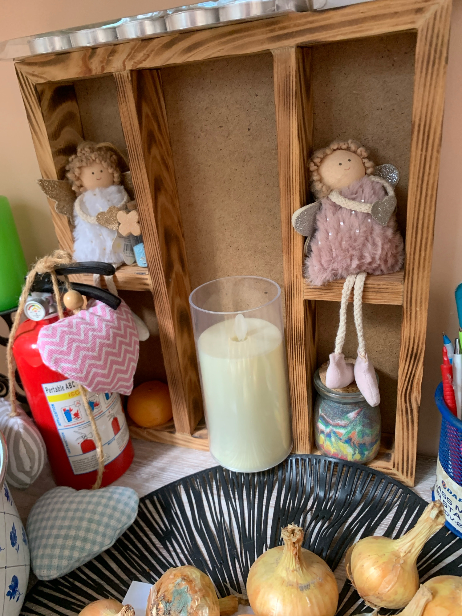 The image displays a wooden shelf unit with two compartments. On the left shelf, a stuffed angel doll with blond curly hair and white clothing is placed. Next to it is a small red fire extinguisher adorned with a pink and white patterned heart-shaped fabric. A single orange fruit sits on the right shelf. On the right compartment of the shelf unit, another stuffed angel doll with blond curly hair, dressed in a pink fuzzy outfit, is positioned. This doll's legs dangle down to a small glass jar filled with colorful sand. In front of the shelf unit, a tall white candle in a clear glass cylinder stands on a black decorative placemat. Below the placemat, three onions are visible, one of which shows signs of spoilage. In the background, a green object and a blue and white patterned fabric heart are partially visible. A blue container holding pens and pencils is situated on the far right. The scene appears to be an indoor display, possibly in a home or shop, with a plain wall as the backdrop. There is no indication of the city of Minsk, Belarus.