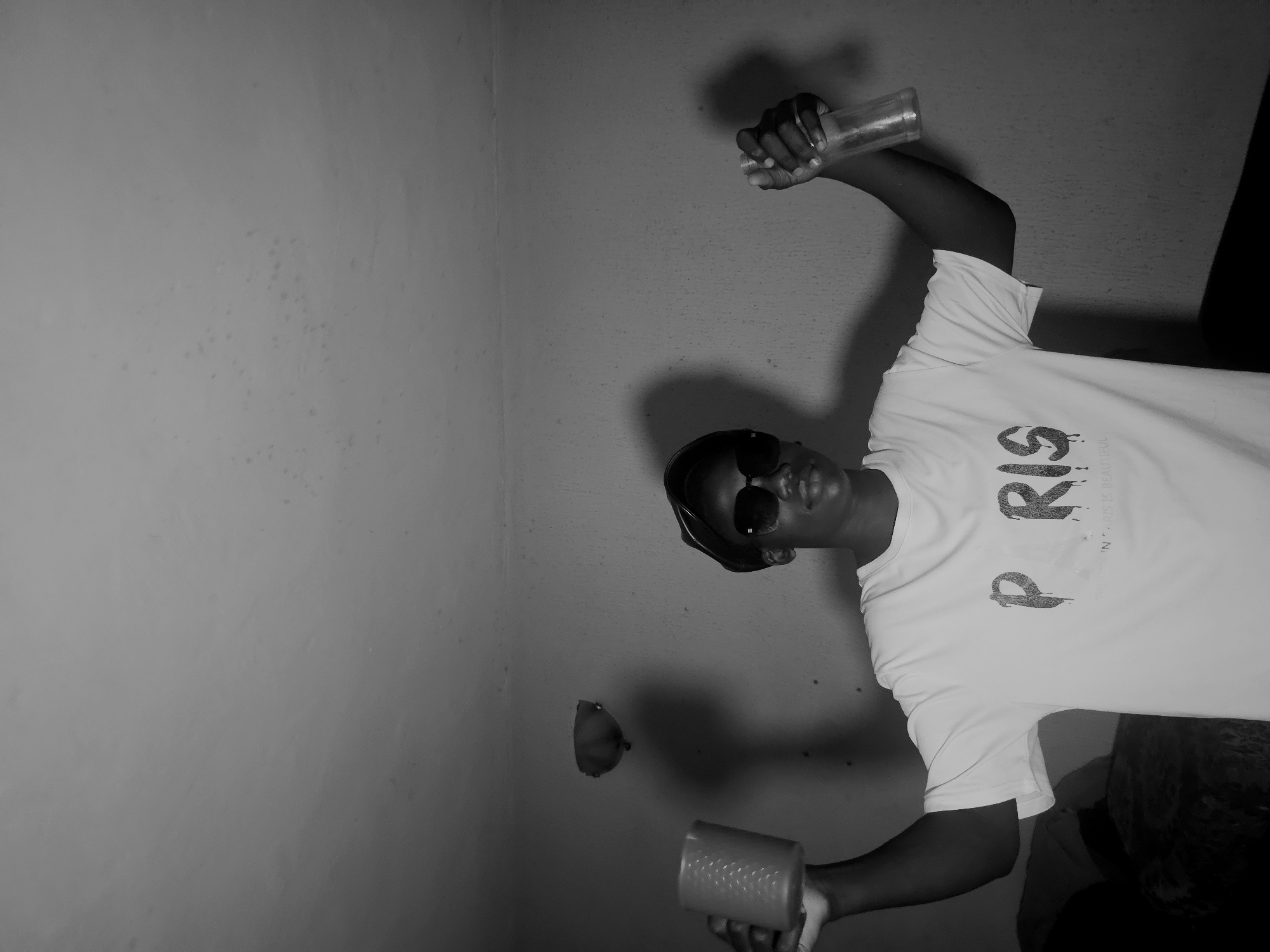 A black and white photograph captures a young person posing indoors against a plain wall in Asaba, Nigeria. The subject, likely male, is wearing a white t-shirt, dark sunglasses, and a dark cap or hat. Their gaze is directed towards the camera.
The white t-shirt features prominent dark text; the most clearly visible part reads "P RIS". Below this, smaller, partially obscured text can be seen, appearing to include words like "IS REAL".
In their right hand, raised above their head, the person holds a clear, cylindrical bottle or glass. Their left hand, held lower, grips a textured cup or glass. The person's body is slightly angled, creating a dynamic pose.
The background consists of a light grey wall with some texture and minor blemishes. Distinct shadows are cast on the wall behind the person, suggesting a direct light source. A small, roundish object, resembling a lemon or a similar fruit, is visible on the wall to the left of the person's head. The image is monochrome, emphasizing contrasts in light and shadow.