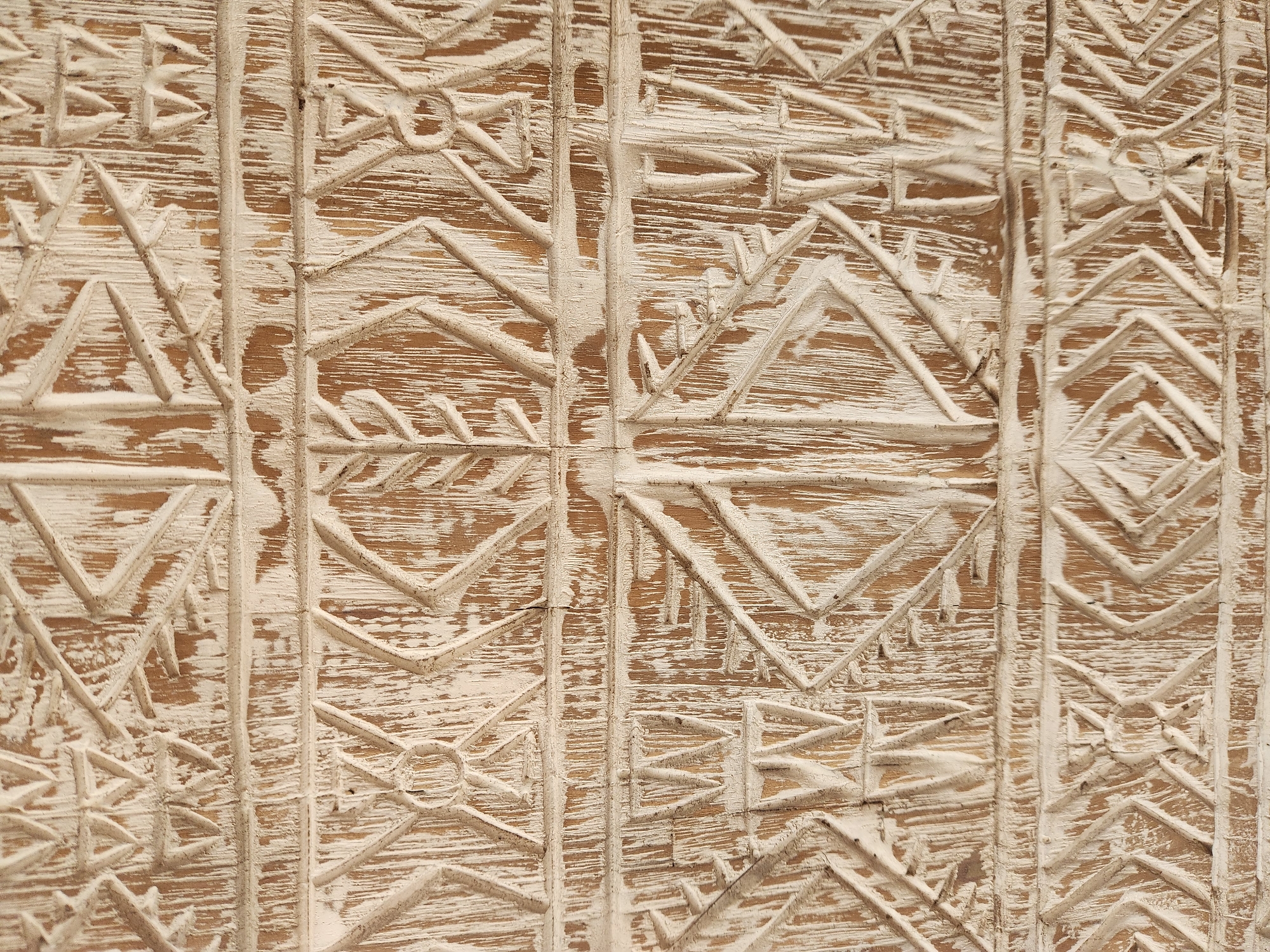 The image shows a close-up, detailed view of a textured, carved wooden surface with an aged, distressed finish. The wood has a light brown, natural grain visible through a white, somewhat chipped or distressed paint. The carvings are geometric and appear to be in relief, forming repeating patterns. These patterns consist of lines, triangles, diamonds, and some circular motifs, creating a sense of depth and texture. The overall impression is of an antique or rustic decorative element. The image does not provide any information about a specific location in Maitland, United States of America.