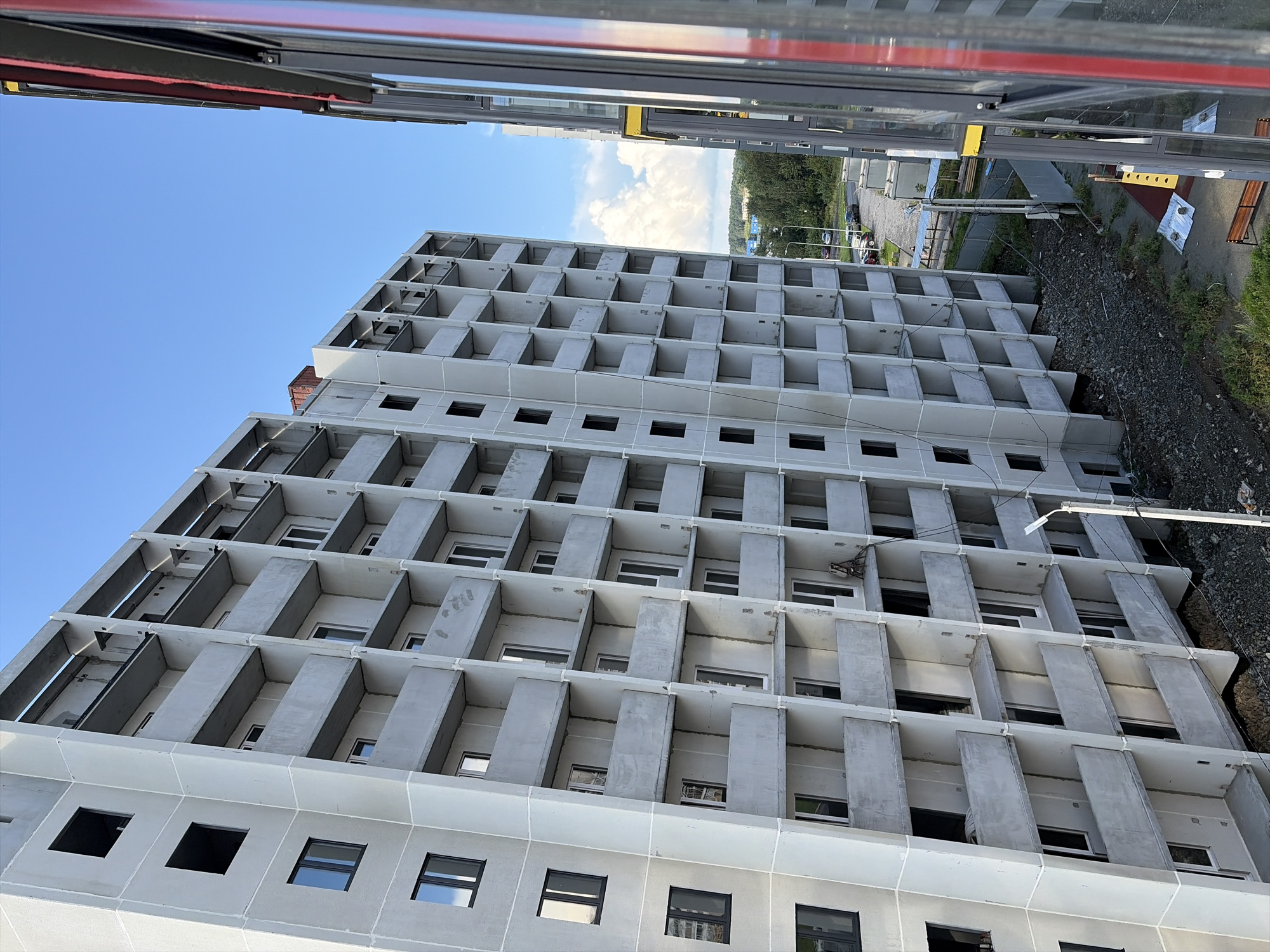 The image depicts the exterior of a multi-story residential building under construction, likely in Yekaterinburg, Russia, given the context. The building is made of concrete, with repetitive balconies and window openings. The facade is unfinished, with exposed concrete and no signs of external finishing. The sky above is blue with scattered clouds. To the right of the building, another structure, possibly a residential building or part of the same complex, is partially visible, featuring modern windows with red trim. The ground area around the construction site is rough, with loose gravel and some scattered debris. In the background, a street with parked cars and trees is visible, suggesting the construction is in an urban or suburban area. A lamppost stands near the building. The overall scene portrays an active construction site.
