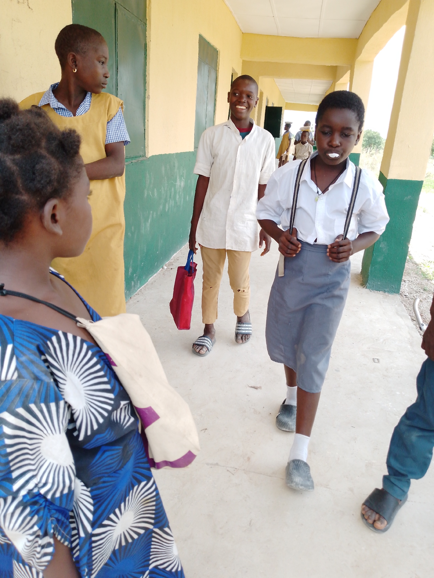 The image shows a group of school children in Akwanga, Nigeria, standing in a covered walkway. A girl in the foreground is wearing a blue and white patterned dress and a light-colored bag. To her right, a girl in a yellow dress and blue checkered shirt looks to the side. Behind them, a boy with a white shirt and khaki pants carries a red bag and smiles. In the center of the image, a boy in a white shirt with suspenders and a grey skirt or shorts walks forward. Other figures are visible further down the walkway. The walkway is painted with yellow above and green below and is open to the outside.