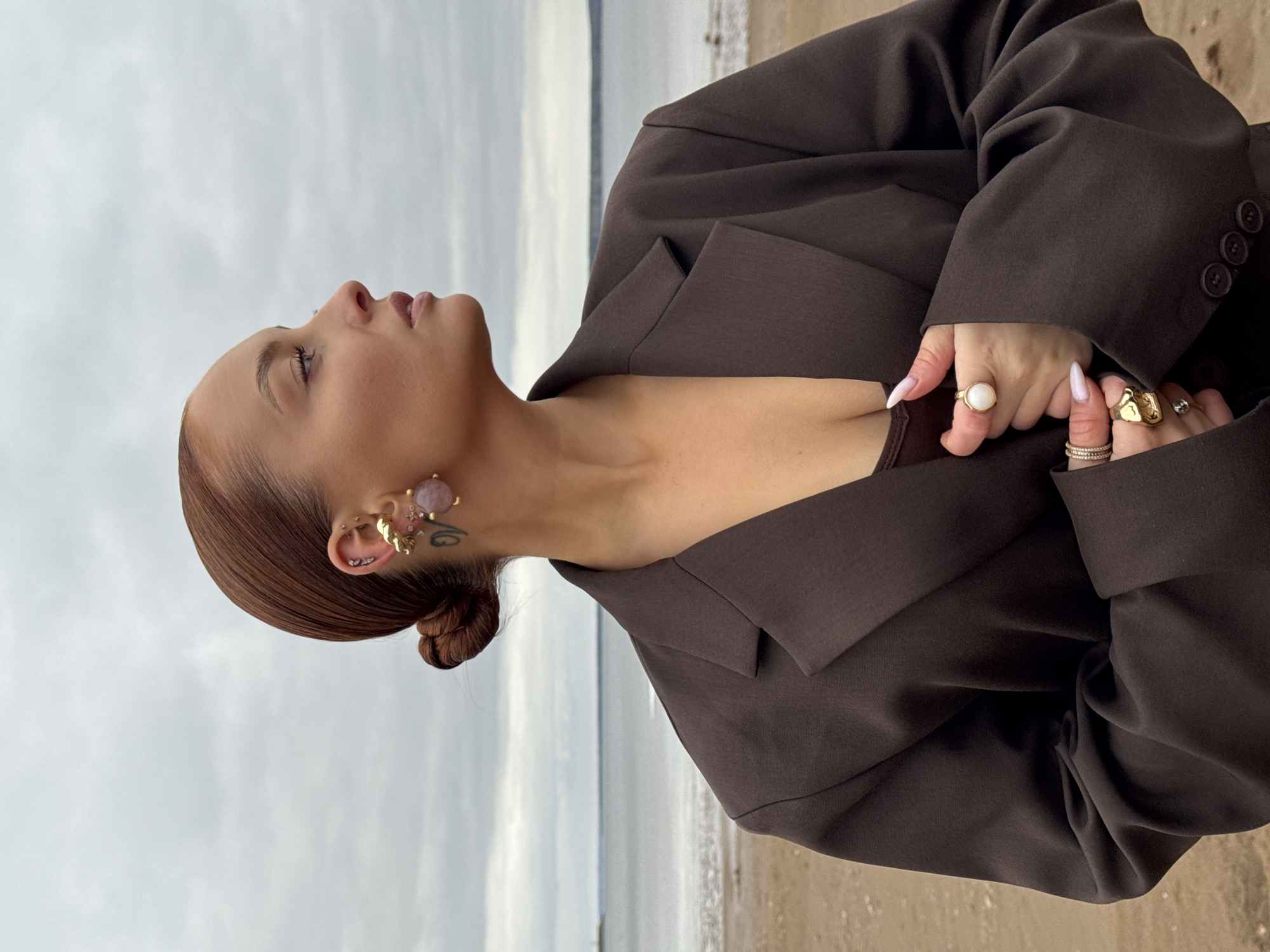 An outdoor portrait captures a person, likely a woman, in a serene pose against a natural backdrop. The subject is shown from the upper chest upwards, viewed in profile, with her head tilted slightly back as she looks upward towards an overcast sky. Her hair, styled in a neat bun at the nape of her neck, is a reddish-brown color.
She is wearing a dark brown or charcoal oversized blazer, which appears to be layered over a matching top or bra. Her right hand is positioned near her chest, gently holding the fabric of her blazer. Her fingers display long, light-colored manicured nails and are adorned with multiple rings; one prominent ring on her index finger features a large, light-colored, possibly opalescent, stone, while other fingers wear gold-toned rings of varying designs. Intricate earrings decorate her left ear, including a large, irregular pinkish-purple stone piece, a gold-toned ear cuff, and several small stud piercings. A subtle tattoo is visible on her neck, just behind her ear.
The setting appears to be a shoreline in Chaikovsky, Russia. In the background, a vast body of water, likely the Votkinsk Reservoir, meets a sandy or earthy shore. The sky is uniformly light gray and cloudy, suggesting an overcast day. No specific activity beyond posing for the photograph is evident, and the overall mood conveyed by her upward gaze is contemplative or calm. No visible text is present in the image.